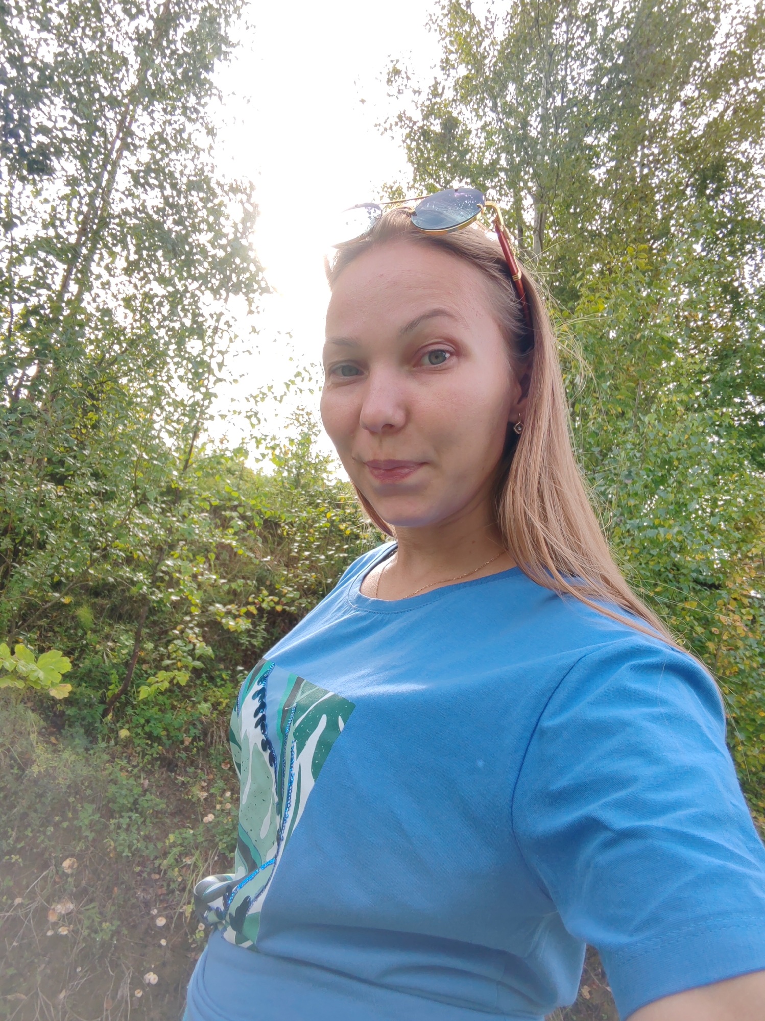 A young woman with blonde hair and sunglasses perched on her head stands in front of a wooded area with the sun shining through the trees. She is wearing a blue t-shirt with a green and white leaf pattern. The location is identified as Chukhlem, Russia.