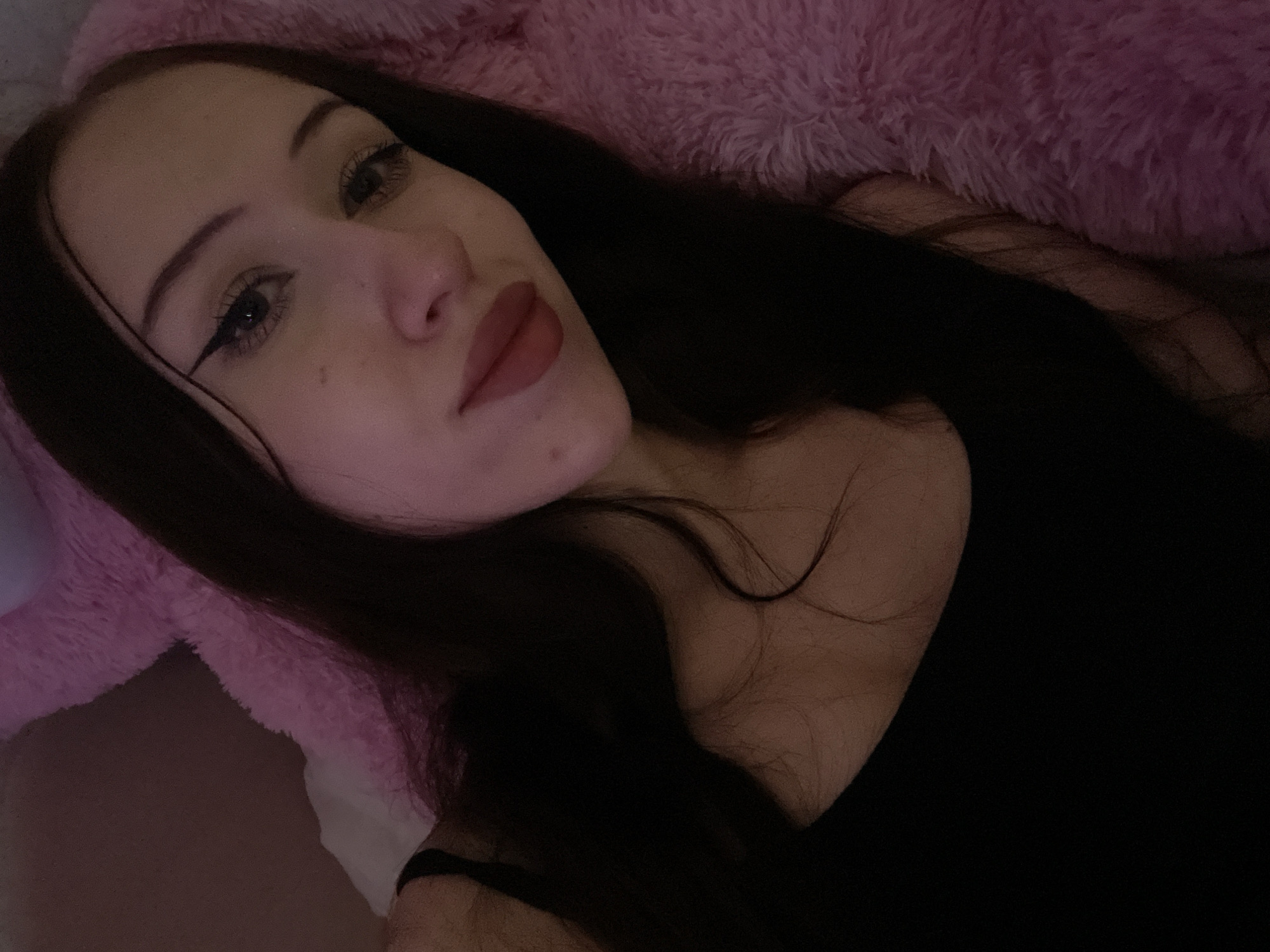 The image shows a young woman with long, dark hair lying down. She is wearing a black tank top and has makeup on, including eyeliner and lipstick. She is against a pink, fluffy surface, possibly a blanket or stuffed animal. The lighting is dim and appears to be artificial, casting a warm glow on her face. There is no indication of the specific location within the city of Votkinsk, Russia, as the image is a close-up and lacks any identifiable landmarks or context.