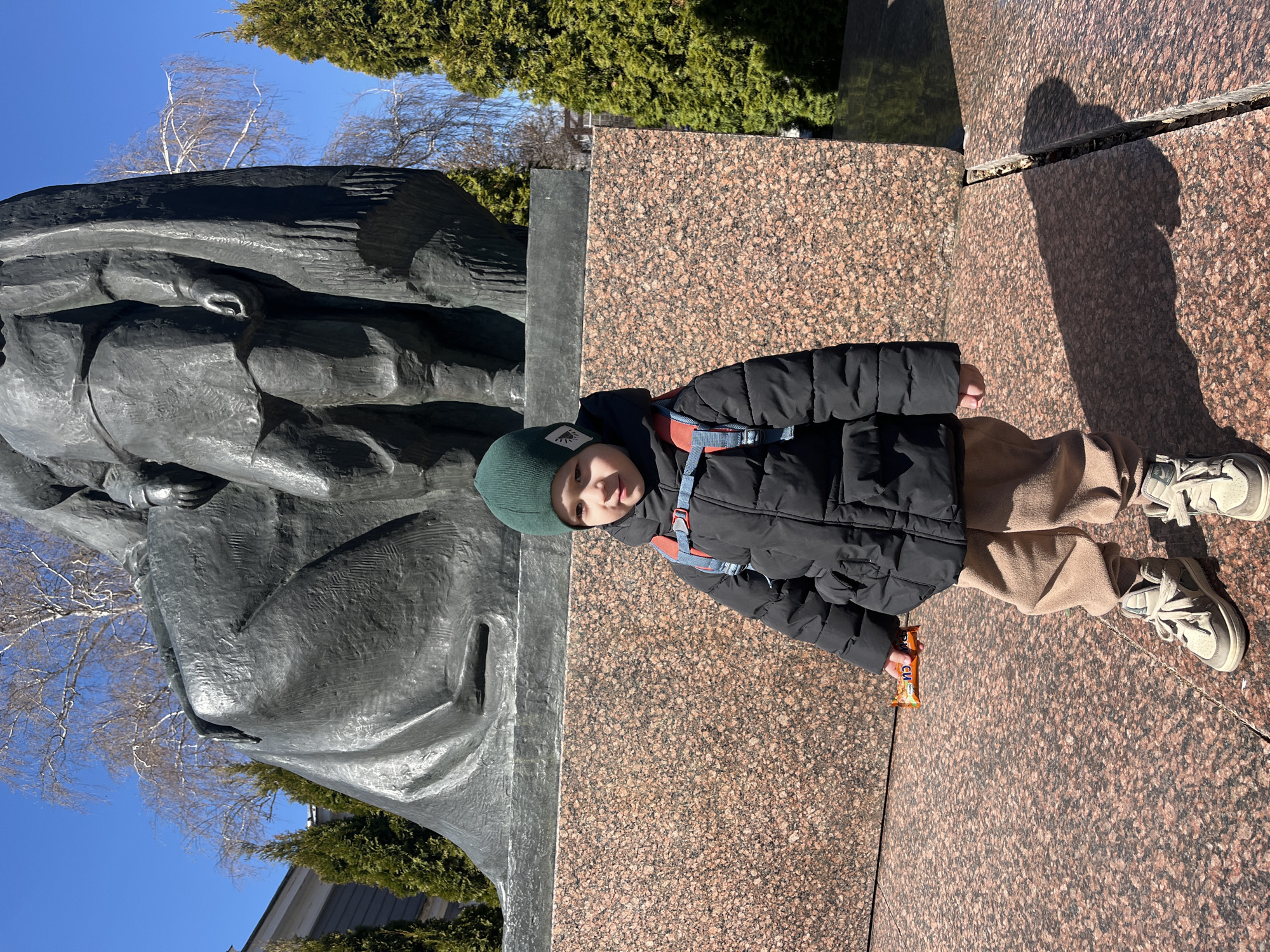 A young boy in a black puffer jacket, tan pants, and grey sneakers is standing on a brown-speckled granite surface next to a large, dark-colored, abstract sculpture. The boy wears a green beanie and has a small snack bar in his hand. The sculpture is positioned on the left side of the image, contrasting with the bright blue sky and bare branches of trees in the background. To the right, behind the boy and sculpture, is a line of evergreen trees. The photograph appears to be taken at an outdoor location. Based on the sculpture style, the scene is likely located at Lenin Memorial in Ulyanovsk, Russia.