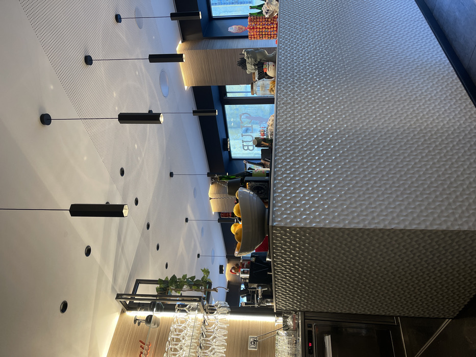 The image displays the interior of a modern bar, café, or lounge, oriented vertically but depicting a horizontal space. A prominent, long bar counter is on the right side of the frame, featuring a distinctive light gray, possibly metallic, hammered or dimpled texture on its front panel.
Above the bar and across the ceiling, numerous black hexagonal pendant lights hang at varying lengths, interspersed with circular recessed lights. Parts of the ceiling also show perforated acoustic panels.
Behind the bar, a variety of items are arranged for service. These include several bottles, a bowl filled with yellow citrus fruits (likely lemons or oranges), and other bar-related equipment. A black multi-tiered shelf below the ceiling further displays potted green plants and rows of inverted clear stemware hanging from racks. In the background, large windows allow natural light to enter the space, and a reflected sign displaying the word "CLUB" (backward) is visible.
No people are present in the scene, suggesting a quiet period in the establishment. The combination of natural light through the windows and the active interior lighting indicates it is daytime. The overall aesthetic is clean, contemporary, and functional, consistent with a modern hospitality venue. No specific emotions are discernible.