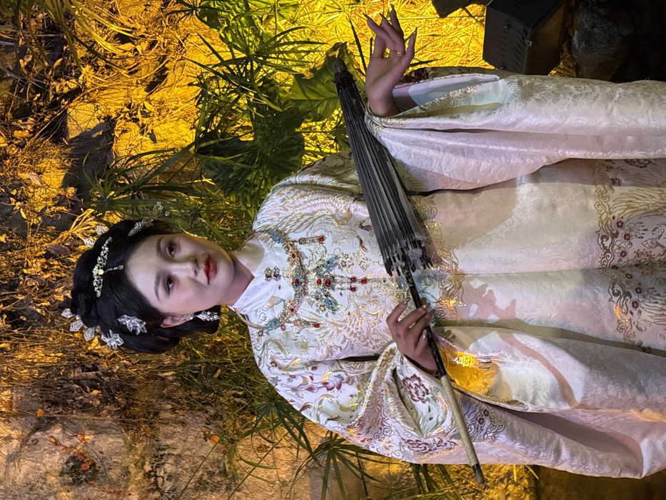 A single East Asian female adult, estimated to be in her 20s or 30s, is centrally positioned in a vertical, waist-up composition, looking slightly left of the viewer with a neutral expression. Her dark hair is styled in an elaborate traditional updo, adorned with numerous silver-toned decorative hairpins, a prominent forehead ornament, and matching earrings. She wears a multi-layered ceremonial necklace featuring blue, gold, and red elements, including red teardrop pendants, over a high-collared white inner garment. Her main attire is a long-sleeved, light-colored (cream or off-white) robe or gown, extensively embroidered with intricate gold and red patterns, consistent with traditional Chinese motifs such as phoenixes and floral designs. Her fingernails are long and manicured. In her left hand, she holds a closed, dark-colored traditional Chinese umbrella diagonally, with the handle pointed downwards. Her right hand is open, fingers slightly spread, with the palm angled towards the viewer. The background features a textured wall or rock face partially obscured by dense foliage, heavily illuminated by strong, warm yellow-orange artificial light, creating distinct highlights and shadows. Large, dark green leaves are visible on the right side of the frame. The scene is located in Yuzhong District, China.