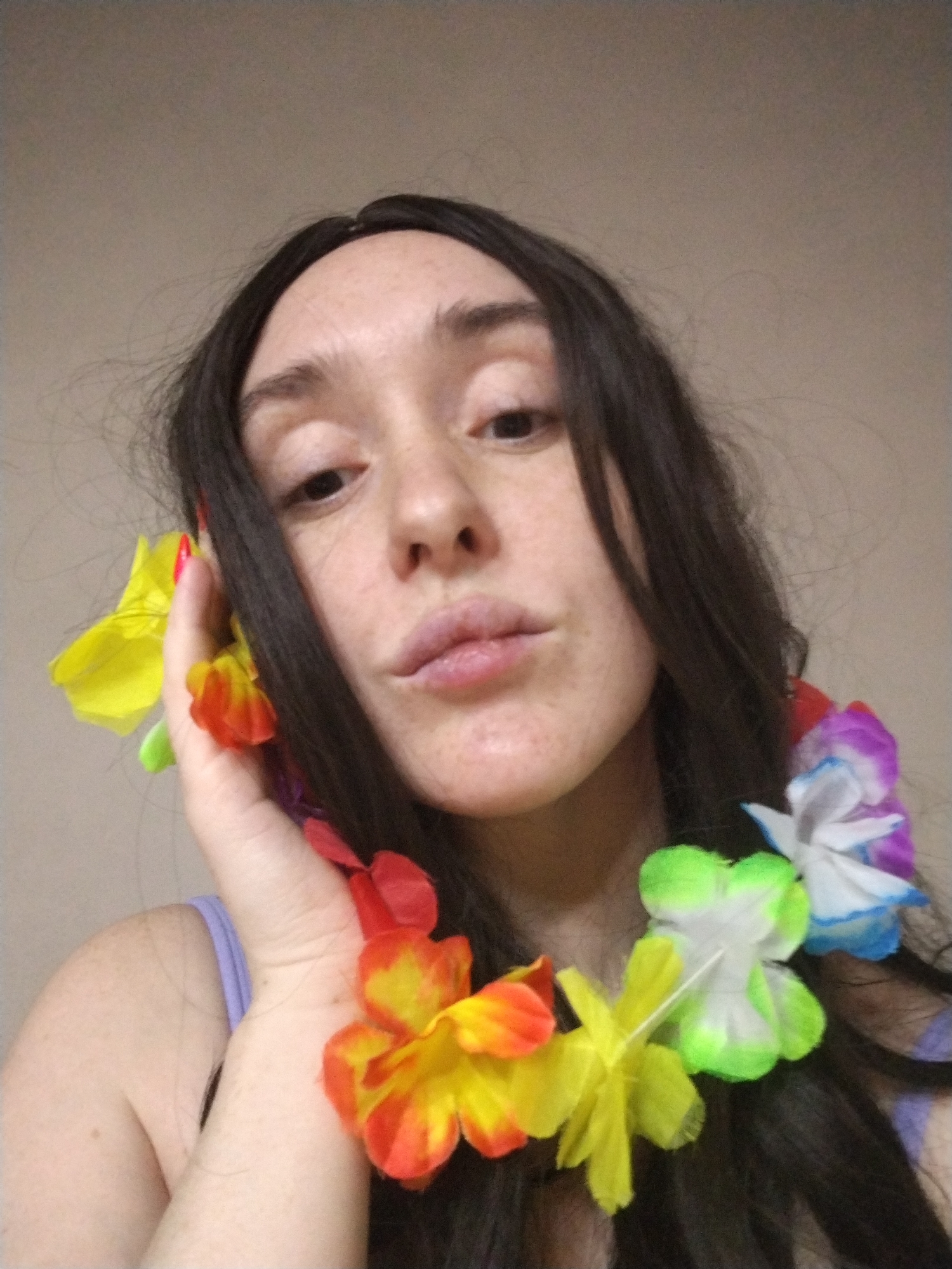 A young woman with dark hair and freckles is posing for a selfie. She is wearing a colorful flower lei around her neck and holding some of the flowers near her face with her left hand. Her lips are pursed slightly. The background is a plain beige wall. There is no indication of the location being Izhevsk, Russia.