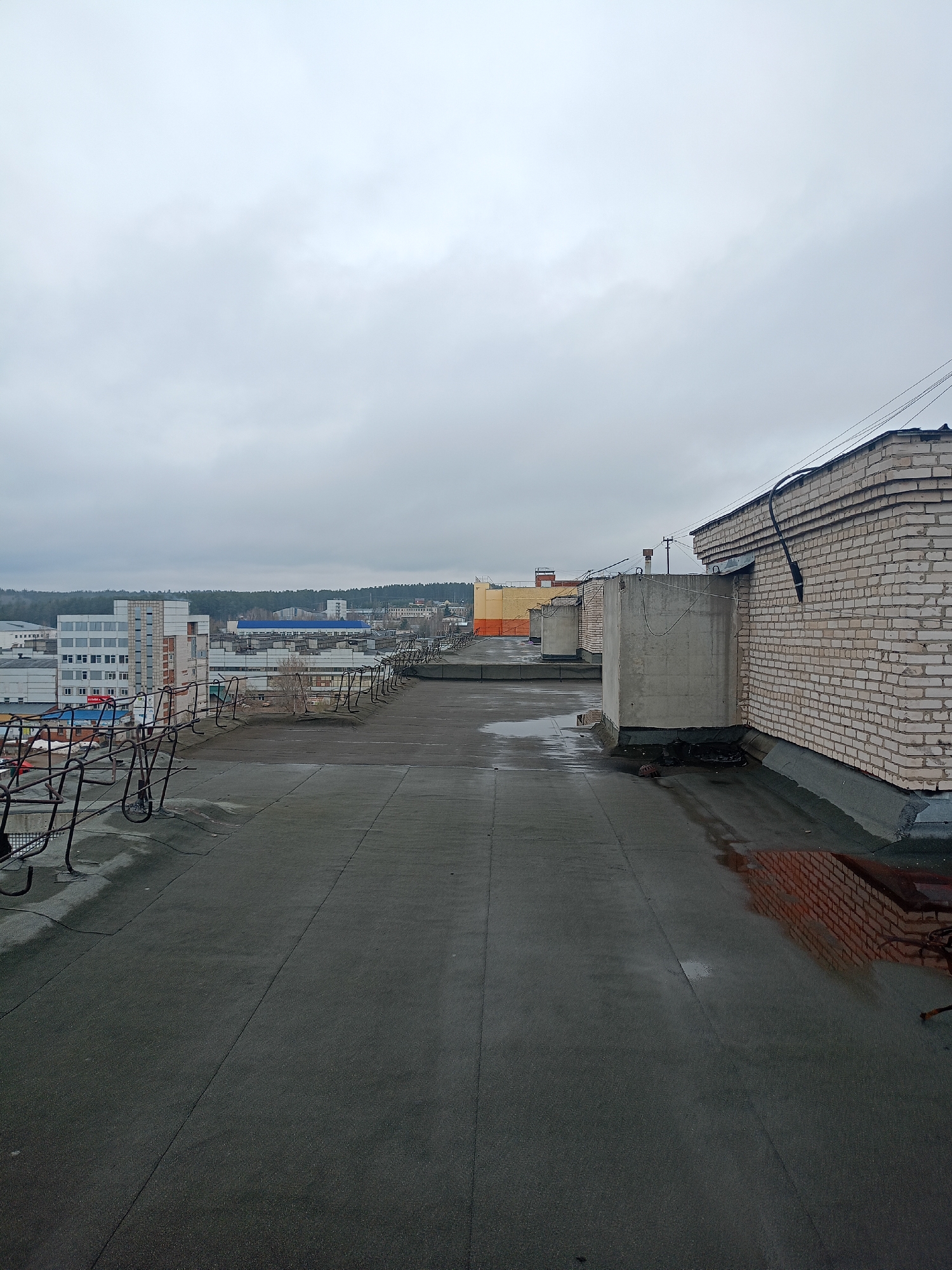 This high-angle image, taken from a flat rooftop in Chaikovsky, Russia, presents an expansive view of an urban landscape under a heavily overcast sky. The foreground is dominated by a dark, possibly asphalt or tarred, rooftop surface that shows wet patches, suggesting recent rain. To the left, a rusty, dark metal railing or fence runs along the edge of the roof, while to the right, a prominent light-colored brick wall of an adjacent building rises, featuring black pipes or conduits and barbed wire along its top.
Further across the immediate rooftop, concrete utility structures are visible, leading the eye towards the broader cityscape. The mid-ground reveals a mix of buildings, including multi-story structures with white facades, industrial-style facilities with blue roofs, and various other architectural forms indicating a diverse urban environment. In the far background, a dense line of trees or a forest stretches across the horizon, providing a natural boundary to the developed area.
The weather is distinctly cloudy, with the entire sky a uniform grey, contributing to a muted and somber atmosphere. It is clearly daytime, though the absence of direct sunlight makes it difficult to pinpoint a specific time beyond broad daylight. There are no people visible in the scene, and no discernible text can be read on any of the structures. The image captures a static, observational moment of an industrial or mixed-use town under overcast conditions.