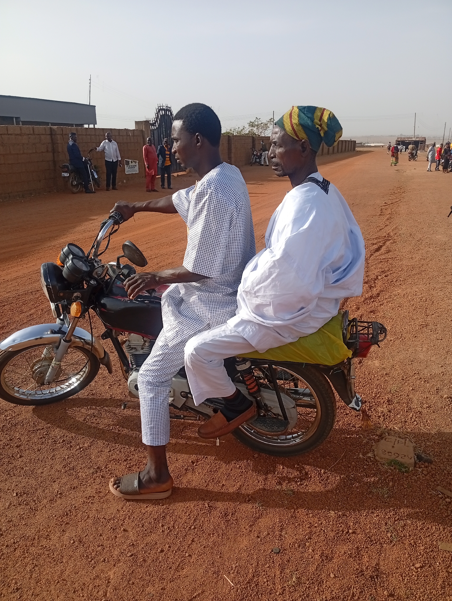 Two men are seated on a motorcycle on a reddish-brown dirt road in what appears to be a semi-urban or rural setting in Du, Nigeria. The man in front, likely the driver, wears a white and blue patterned tunic and trousers, with sandals on his feet. He has his hands on the motorcycle handlebars, looking towards the right side of the frame. The passenger, an older man, is seated behind him, wearing a white flowing robe and a colorful striped cap. He is looking towards the right, similar to the driver.
The motorcycle is black with chrome details, including the front fender, handlebars, and exhaust pipe. A yellow cover or bag is draped over the passenger seat. The ground is dusty, reddish-brown soil with small pebbles. Shadows indicate it is daytime, likely late morning or early afternoon, under a clear, bright sky.
In the background, a low brick wall runs horizontally across the frame, with a black metal gate visible. Several people are standing near this gate and further down the road, some appearing to be conversing or waiting. Another person is seated on a motorcycle near the wall to the left. The overall scene suggests a typical day in a bustling community.
On a small concrete block partially buried in the dirt in the lower right foreground, the text "CPL" and "86929" is visible. On a sign mounted on the wall in the middle background, the number "200" is discernible, followed by partially obscured text.