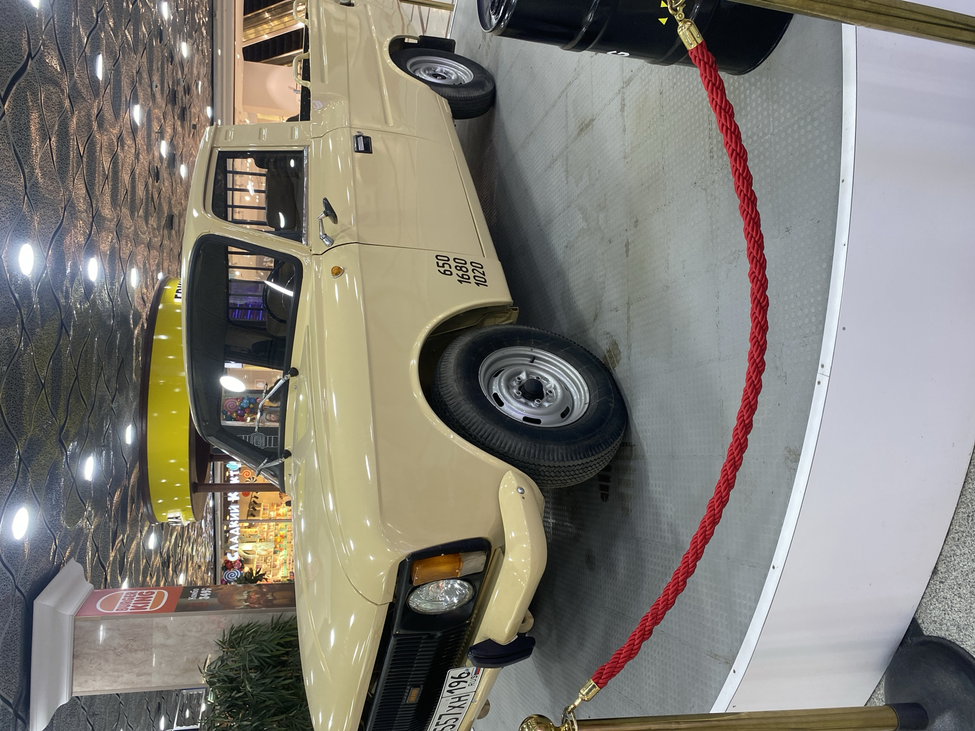 The image shows a light tan-colored vintage truck on display indoors, likely a car show or museum exhibit. The truck is a four-door model, parked on a gray-tiled platform with a white curved edge. It is sectioned off by a red rope barrier with gold-colored stanchions.
The truck has black numbers "650 1680 1020" printed on the side of the truck body, and a license plate "557 XH 196 RUS." The headlights are visible, and the wheels have silver rims.
In the background, there's a shopping center or mall setting, identifiable by storefronts and decorative elements. Specifically, one store sign is visible, written in Cyrillic: "СЛАДКИЙ К КТ," along with a Burger King sign. This suggests that the location is likely in Russia, and the license plate confirms this.
The environment includes a patterned wall in the background, likely part of the mall's design. The lighting is artificial, creating reflections on the vehicle's surface. Given the mall's presence and the objects within it, the specific location is most likely Yekaterinburg, Russia.
