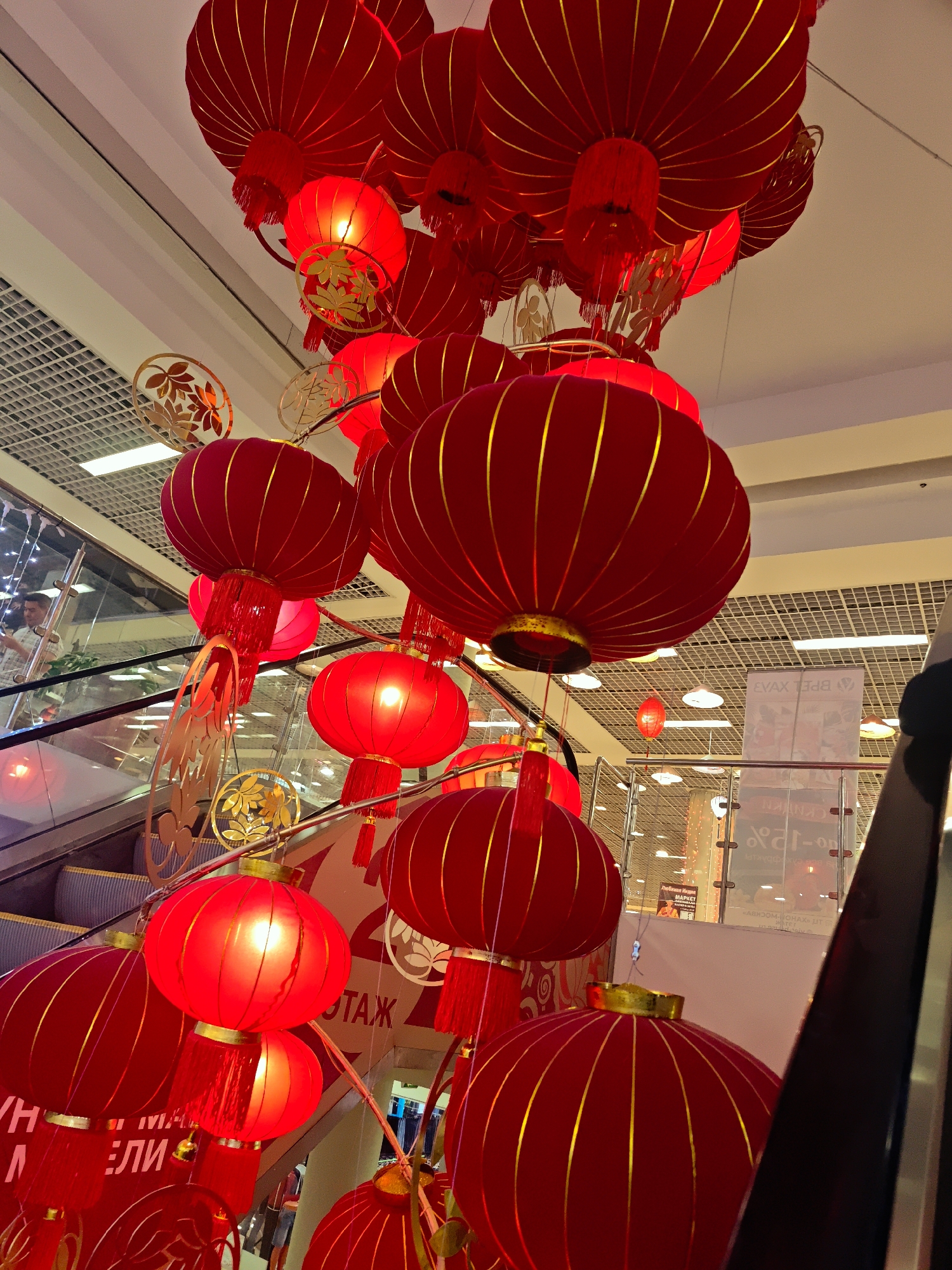 This upward-angled, full-frame shot captures a festive indoor scene adorned with numerous red and gold Chinese lanterns, likely celebrating Lunar New Year or a cultural event, within a shopping mall or large commercial building in Moscow, Russia. The lanterns, varying in size and shape, hang prominently from the ceiling, many illuminated and casting a warm red glow. They feature golden vertical stripes and decorative tassels, with some smaller, gold-colored leaf or floral-patterned ornaments interspersed among them.
An escalator is visible on the left, with a man ascending while looking at his phone. His reflection can be faintly seen in the adjacent glass railing. Another escalator is partially visible on the far right. The ceiling above features a grid pattern with integrated lighting.
Several pieces of Cyrillic text are visible on signs and as reflections. The word "ЭТАЖ" (meaning "Floor") is clear on a sign to the left. Other partial Cyrillic words such as "ЛИ" and "MAPKET" (Market) are discernible. Inverted and reflected text on the right includes "BPEL XVAR" and "«ABDOM-OHAX» UT", along with "-12" and "CKNYKN", confirming a commercial setting in Russia.