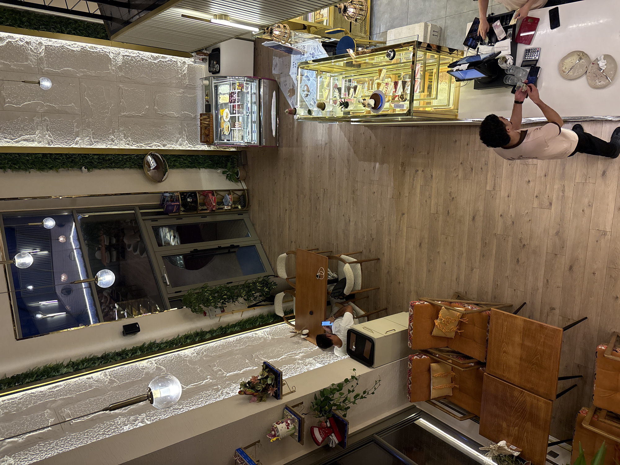 This is an aerial view of the interior of a cafe, likely located in Khatay Raion, Azerbaijan. The cafe has a modern design with wooden floors and white textured walls adorned with vertical green plant installations and accent lighting.
In the foreground, two individuals are interacting at a counter. One person, dressed in a light-colored t-shirt and black pants, is facing the counter and appears to be handling money or receipts. Another person, partially visible, is also at the counter and seems to be working. The counter features a POS system with two monitors, a calculator, and various items like glasses and plates. Display cases filled with pastries and desserts are visible behind the counter.
Further into the cafe, a refrigerated display case is filled with colorful desserts and ice cream. Seating areas are visible, including a dining table with chairs where a person is seated and looking at a phone. Smaller seating arrangements with orange chairs and tables are also present.
Decorative elements such as hanging pendant lights, shelves with decorative items, and potted plants contribute to the ambiance of the cafe. The overall scene depicts a functioning cafe environment with customers and staff present.