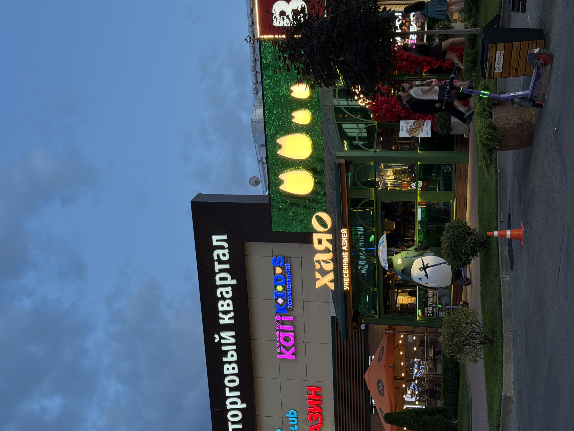 This is a vertical, eye-level, outdoor shot of a commercial building with signage and people. The time of day appears to be dusk, with a twilight sky and soft lighting emanating from the building. The building has a modern facade with large glass windows and prominent signage.
The most prominent sign, in large white letters, reads "Торговый квартал" which translates to "Shopping Quarter." Below this, there are other signs in various colors and languages, including "Kari Kids" and "Хаяо" which seems to be a name. One sign also mentions "ЗАВТРАКИ" (Breakfast) and "УНЕСЕННЫЕ АЗИЕЙ" (Taken to Asia), suggesting the presence of food establishments.
Several people are visible in the scene, some walking and others gathered near the entrance. One individual is captured in motion, appearing to be walking. Another group is standing near a trash receptacle and a scooter. The building's green facade is decorated with glowing yellow shapes that resemble cat ears, adding a whimsical touch.
In the foreground, a street runs alongside the building, with a paved sidewalk and a strip of greenery. An orange traffic cone is visible on the pavement. The overall atmosphere suggests a bustling urban commercial area at the end of the day, possibly a shopping center or a street with various businesses. The presence of scooters hints at a modern, active urban environment.