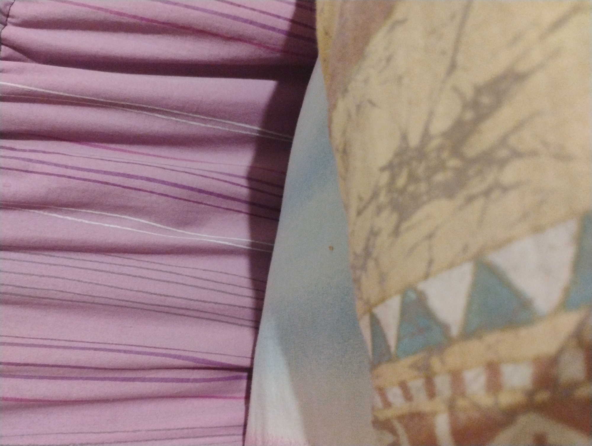 A close-up, overhead shot shows a bed with striped bedding and a pillow. The bedding is a light purple with thin vertical stripes in shades of purple and white. The pillow is light blue and rests against the bedding. In the foreground, a comforter with a geometric pattern in shades of tan, brown, white, and blue is visible. The image is of an interior scene, likely a bedroom, with no specific indicators of its location in Plachkovtsi, Bulgaria.