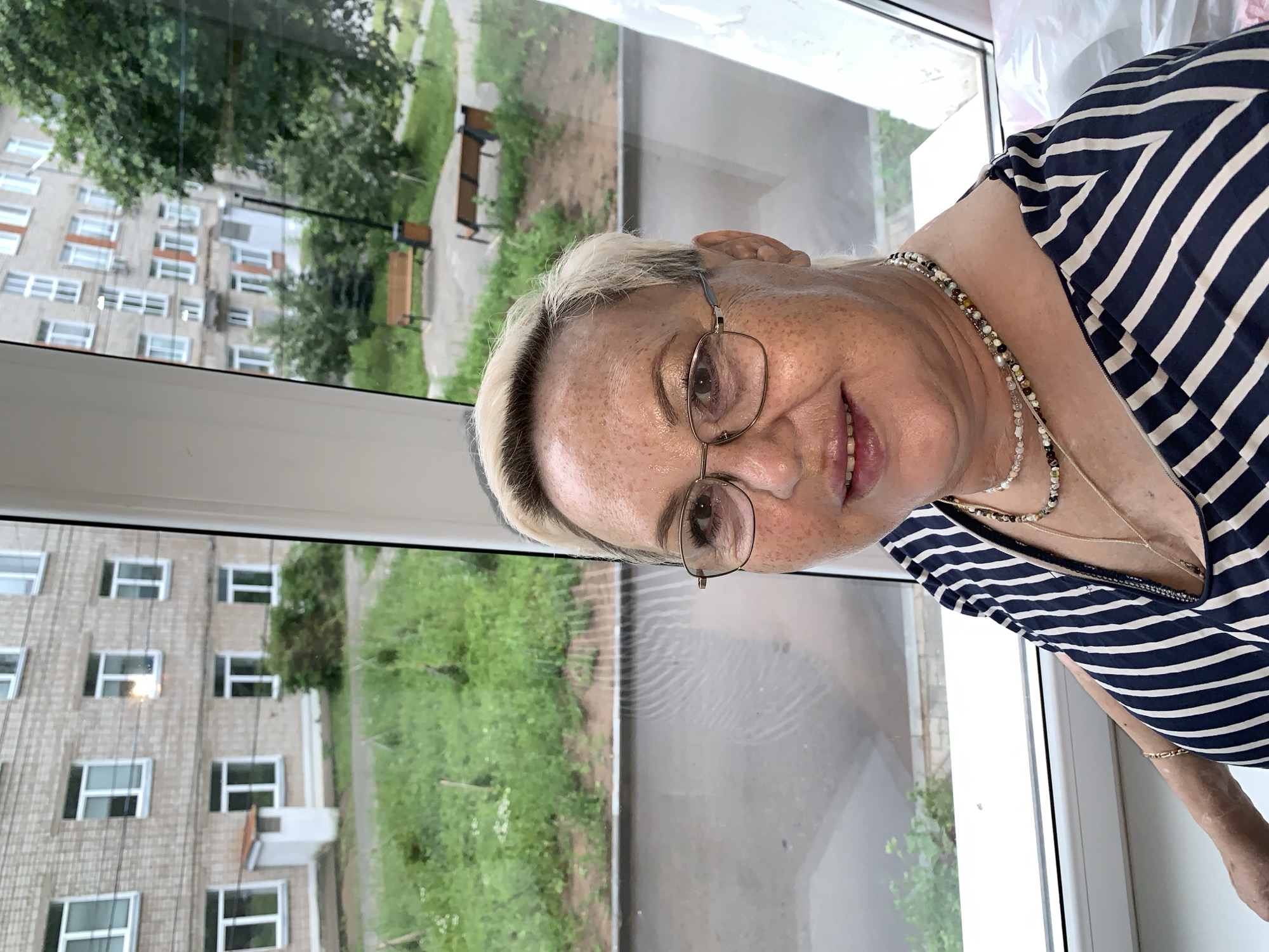 A medium shot portrait shows a woman with short, blonde hair, freckled skin, and glasses. She is wearing a dark blue and white striped top and several necklaces. The woman is positioned in front of a large window, which offers a view of an apartment building and an outdoor courtyard with green grass, trees, and park benches. The interior of the room is visible in the foreground, with a white windowsill and the corner of a plastic bag. The exterior view through the window suggests a setting in a city like Chaikovsky, Russia, with typical Soviet-era apartment blocks and a communal green space. The lighting is natural, coming from the window.