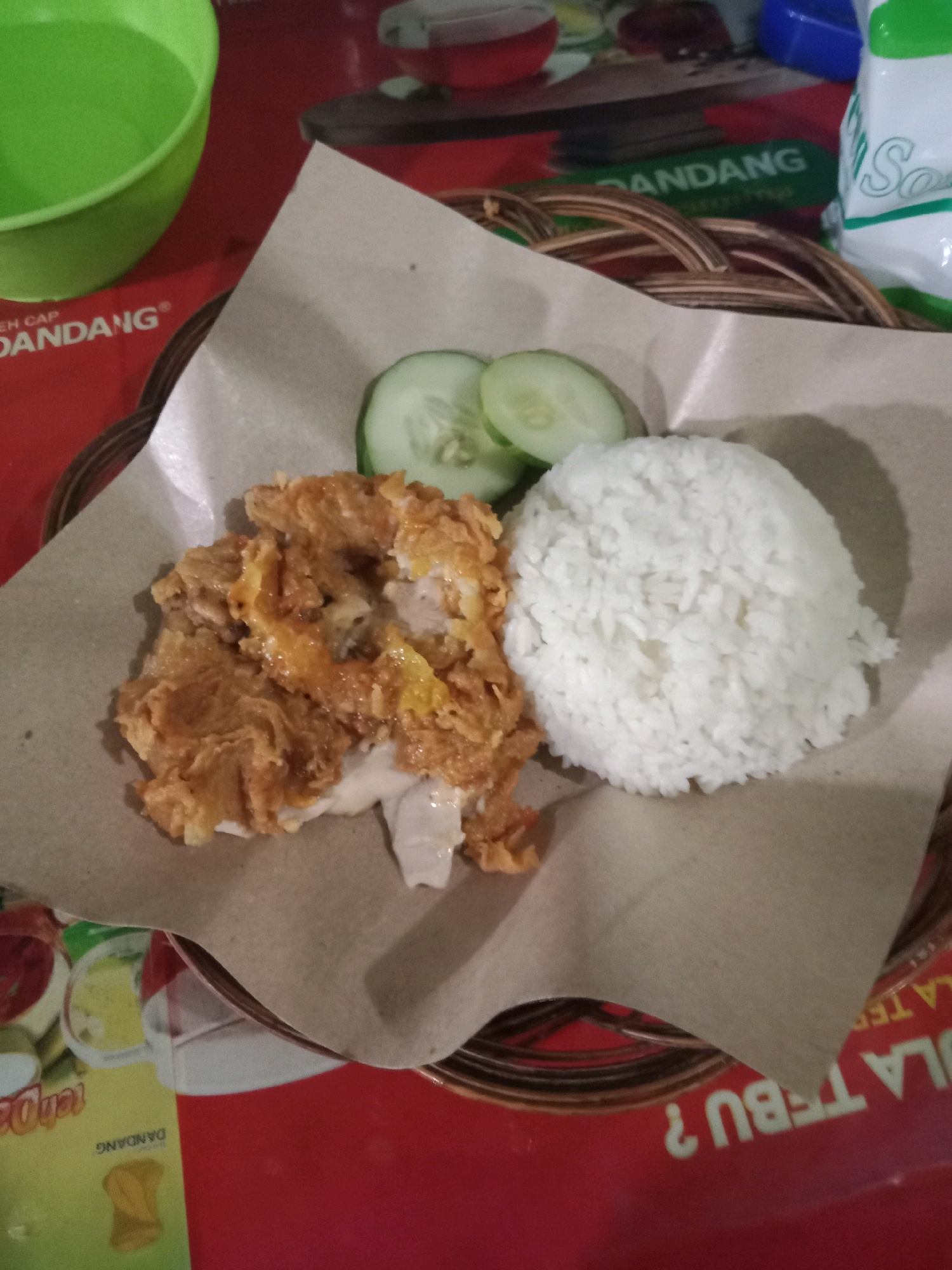 The image shows a close-up overhead view of a plate of Indonesian food, likely served in a casual eatery or street food stall in Semarang, Indonesia. The meal consists of a portion of fried chicken, a mound of white rice, and three slices of cucumber, all arranged on brown paper lining a woven rattan plate. The fried chicken appears golden brown and crispy, with visible pieces of meat. The rice is fluffy and white. The cucumber slices are fresh and green. The background shows a red surface, possibly a table, with glimpses of other items, including a green bowl and what appears to be packaging for food products. The lighting suggests an indoor or shaded outdoor setting. The overall scene conveys a simple, everyday dining experience.