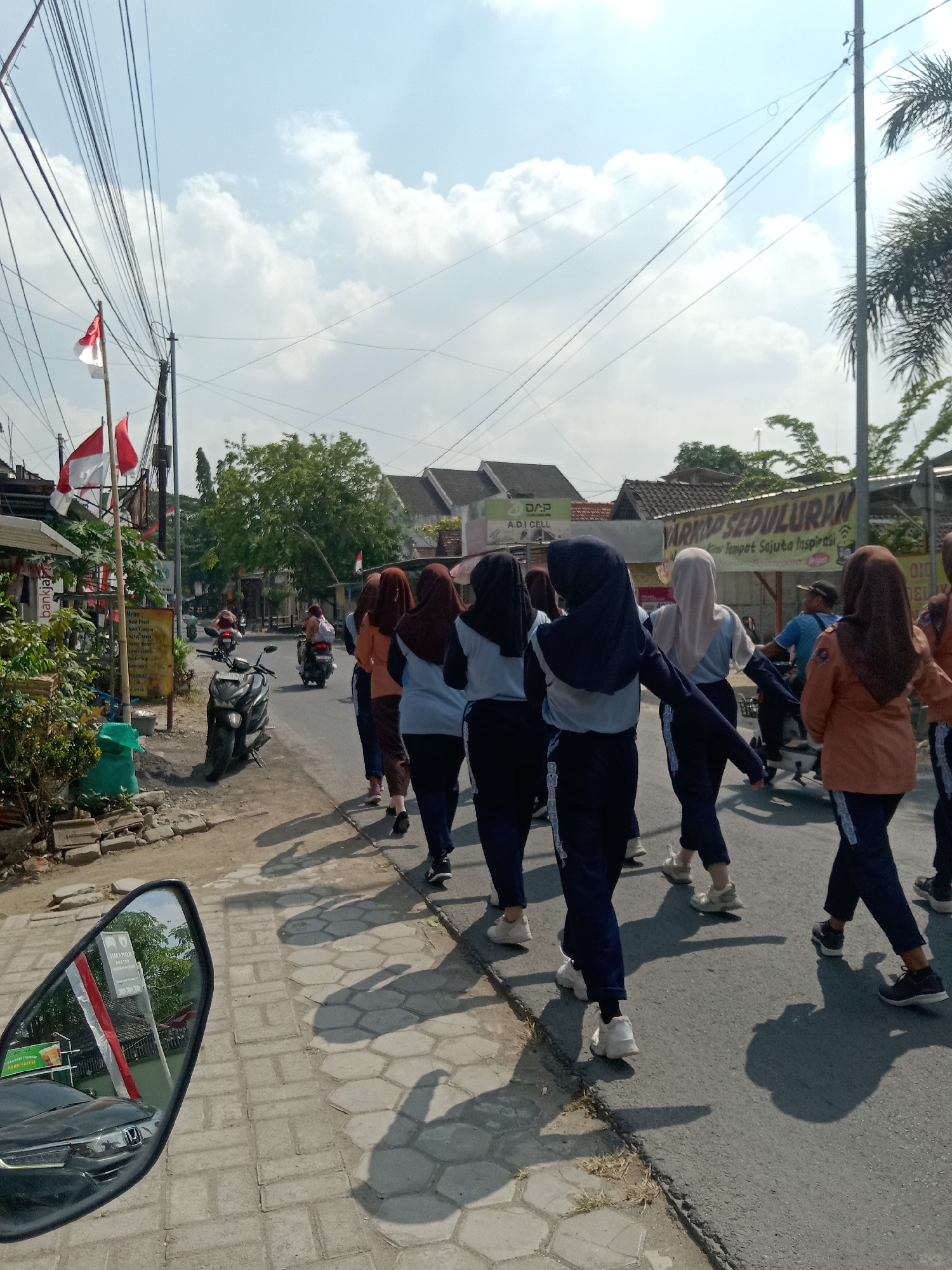A group of young people, dressed in what appears to be a school uniform consisting of dark blue pants and light blue shirts, are walking along a paved road. Some are wearing hijabs in various colors including dark blue, light blue, and brown. They are walking in a single file line away from the camera. In the background, there are several buildings, trees, and a clear blue sky with some clouds. Two red and white flags, possibly of Indonesia, are visible on poles on the left side of the road. A black scooter is parked on the left side of the road. The road is lined with hexagonal pavers on the left side, and the right side has a darker asphalt surface. Shadows of the people walking are cast on the road. A side mirror of a vehicle is visible in the foreground on the bottom left, reflecting a car and the flags. There are also signs in Indonesian visible on buildings in the background, suggesting an urban or semi-urban setting in Indonesia, likely Sugihwaras, Indonesia given the context. A person is riding a motorcycle in the distance.