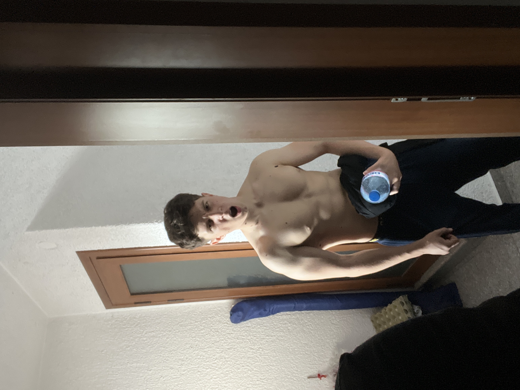 An indoor, vertical medium shot depicts a young adult male with a muscular build, bare-chested, and wearing dark trousers. He has short, dark, curly hair and a pale complexion. His mouth is open in an apparent yell or exclamation, with a furrowed brow and wide eyes, looking directly towards the camera. He holds a clear plastic water bottle with a blue cap and label in his right hand, partially obscured by dark fabric draped over his arm. His left arm hangs naturally by his side. The background features a white, textured wall on the left, against which a blue rolled object (possibly a mat) and a white egg carton are visible. A light brown wooden door with an opaque glass panel is positioned behind the man's left side. To the right of the man, a dark brown wooden door or wardrobe is partially visible. The floor is a light-colored tile. The lighting is uniform, suggesting an indoor setting. The scene is located in the city of Unknown, Unknown.