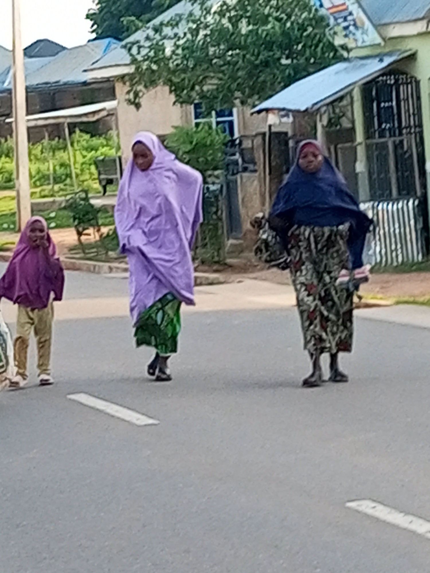 The image depicts a street scene in Tempure, Nigeria. Three individuals are visible, walking on a paved road. The person on the left is a child wearing a purple head covering and tan trousers. To their right is an adult wearing a light purple, flowing garment and a green patterned skirt. The third individual, on the far right, is an adult wearing a dark blue head covering and a patterned skirt. Buildings with corrugated metal roofs and some vegetation are visible in the background. The lighting suggests it is daytime.
