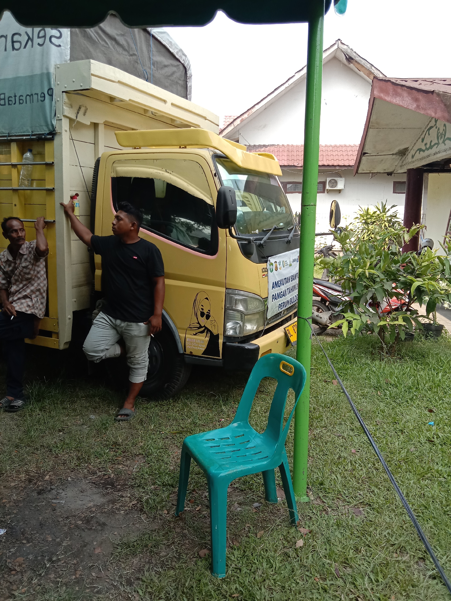 The image depicts an outdoor scene in Lhokseumawe, Indonesia. Two men are near a yellow truck. One man, dressed in a plaid shirt, leans against the truck's side. The other man wears a black t-shirt and grey pants, leaning on the truck's front. A banner on the truck's front reads "ANGKUTAN BANTU PANGAN TAHUN 2 PERUM BULO." A green plastic chair sits on the grass next to a green pole. In the background is a white building with a red roof, and motorcycles are parked nearby. The ground is covered in grass.