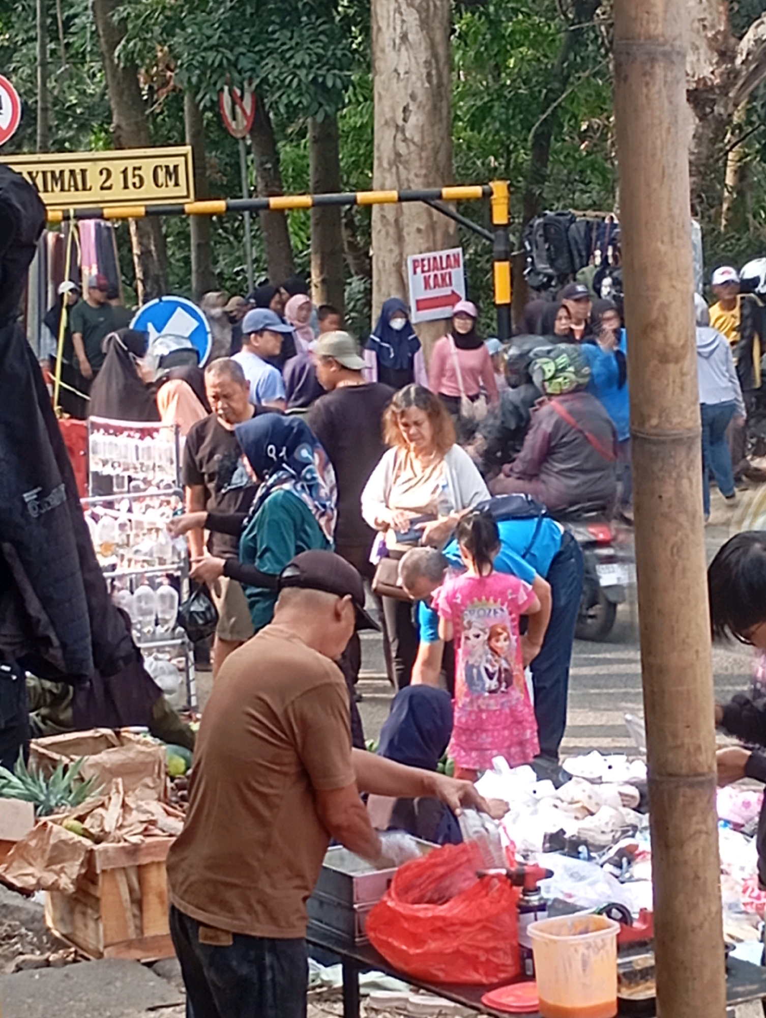 The image depicts a street scene in Cimahi, Indonesia, likely an outdoor market or roadside vendors. A yellow and black striped barrier with signage is visible, including a sign indicating a maximum height of 2.15 meters. Another sign with red text and an arrow points to "PEJALAN KAKI," meaning pedestrian. People of various ages are present, some browsing goods displayed on tables and stalls, while others are walking or standing. Among the items for sale are what appear to be small packaged goods, possibly food items or drinks in clear plastic containers, arranged on shelves. A wooden crate containing pineapples and other produce is also visible. A young girl in a pink dress featuring cartoon characters is a prominent figure in the foreground, interacting with adults. Clothing items, bags, and personal belongings are scattered on the ground and on display. The environment is characterized by trees and a paved road, suggesting an urban or semi-urban setting.