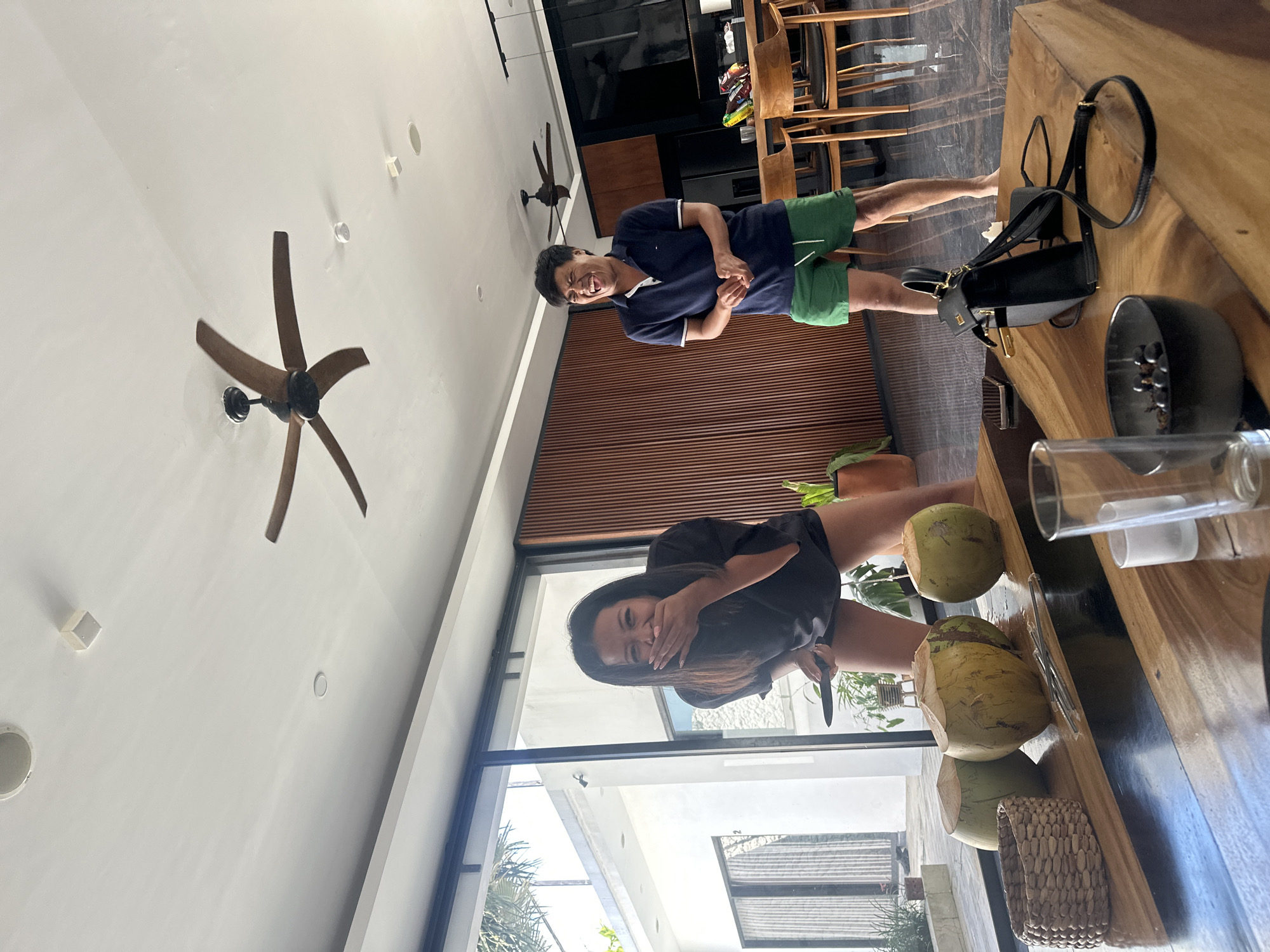 The image shows a candid moment between a man and a woman in a modern, indoor setting that appears to be a living or dining area. The man, wearing a navy blue polo shirt and green shorts, is making a comical face with his mouth open wide and eyes squinted. He is standing near a wall with vertical wooden slats. The woman, wearing a black t-shirt, is seated and covering her mouth with her hand, also appearing amused or surprised.
In the foreground, a large wooden table dominates the frame, with several coconuts, a woven basket, and a glass are visible on its surface. A black handbag with gold accents rests on the table. Behind the table, a ceiling fan with wooden blades is visible. Large windows allow natural light to enter the space, suggesting it is daytime. The overall atmosphere is relaxed and informal, hinting at a casual gathering or a moment of shared humor. The location is identified as North Kuta, Indonesia, which might imply a tropical climate, consistent with the presence of coconuts. No visible text is present in the image.