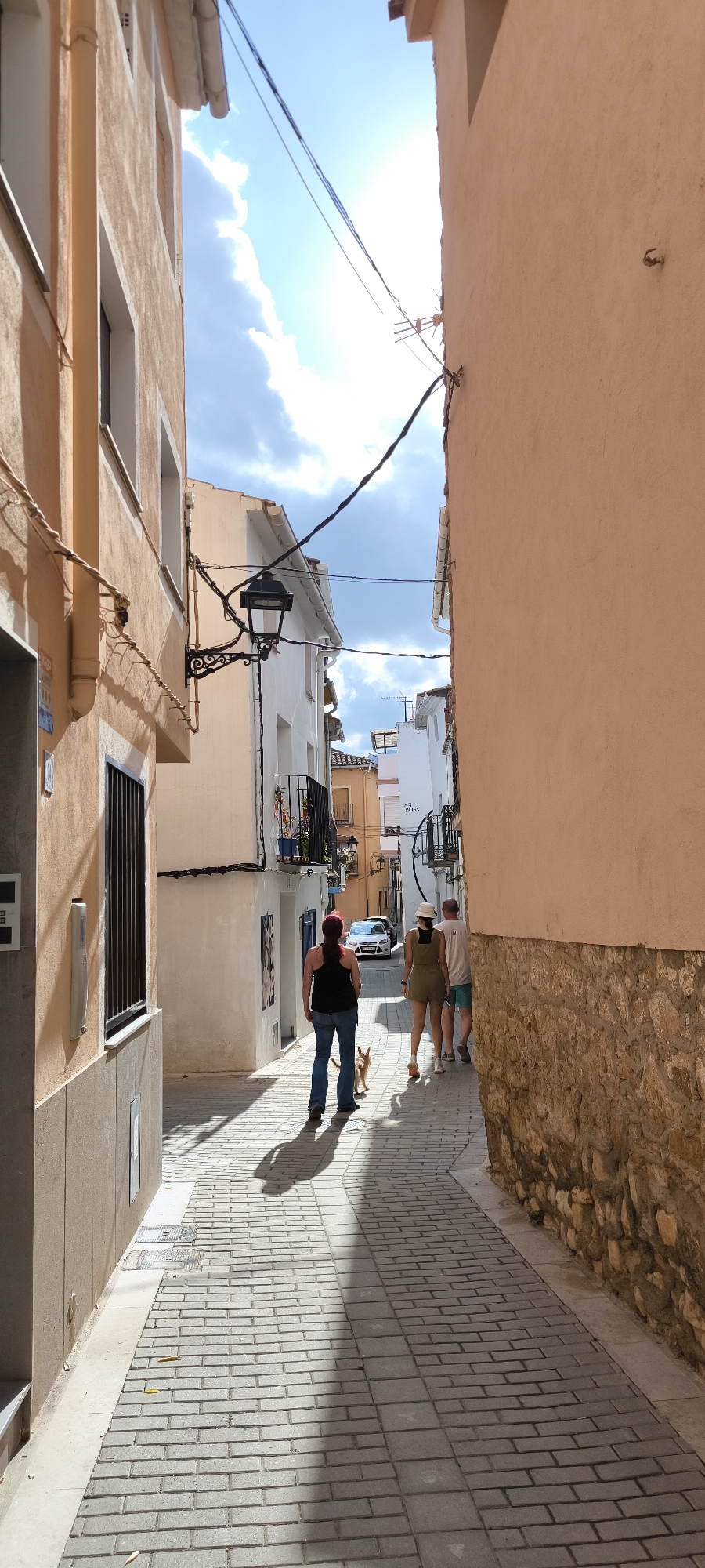 The image depicts a narrow street in Fanzara, Spain, with buildings lining both sides. The street is paved with light-colored bricks, and sunlight casts shadows across it.
In the foreground, a woman with red hair is walking a small, light-colored dog on a leash. She is wearing a dark sleeveless top and blue jeans. Behind her, a couple is walking away from the camera. The woman is wearing a khaki-colored romper and a white hat, while the man is wearing a white t-shirt and blue shorts.
The buildings on the left are a light orange color with white window frames. One building has a dark-colored barred window. A black, ornate street lamp is mounted on the wall of a white building on the left. The building on the right is a light orange color with a rough, stone-like lower section.
Further down the street, a white car is parked. The sky above is blue with white clouds, and power lines are strung across the street. The overall scene suggests a quiet, sunny day in a Mediterranean town.