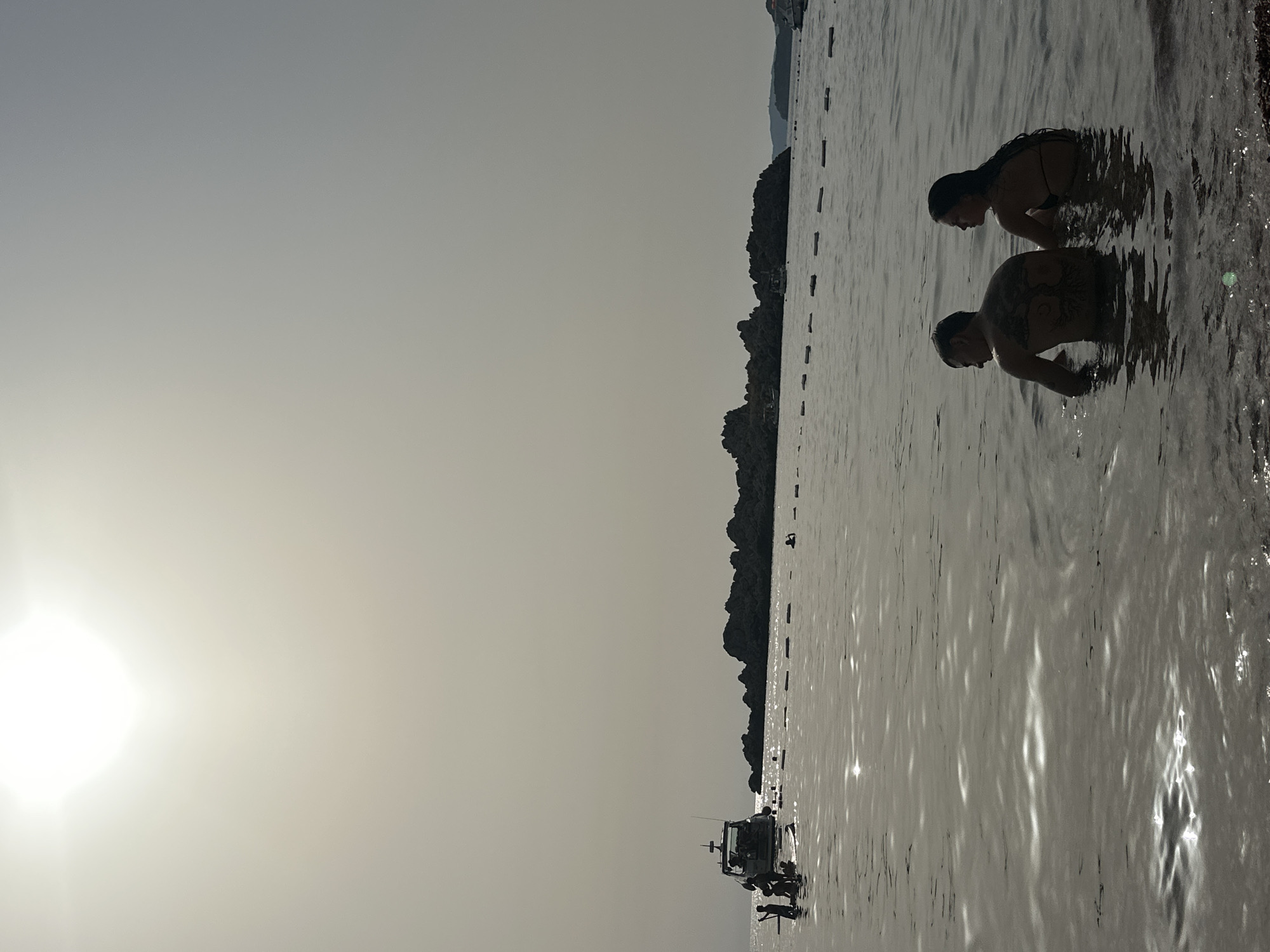 In Sveti Stefan, Montenegro, a man with a tattooed back and a woman with wet hair are partially submerged in calm, reflective water. They are facing away from the camera, appearing to be conversing or looking out at the sea. The sun, high and bright, casts a soft glare across the hazy sky. In the distance, a small boat is anchored near a rocky island formation, with a bu oy line marking off an area of the water. Several figures are visible on and around the boat and in the water further out. The shoreline in the foreground is composed of pebbles.