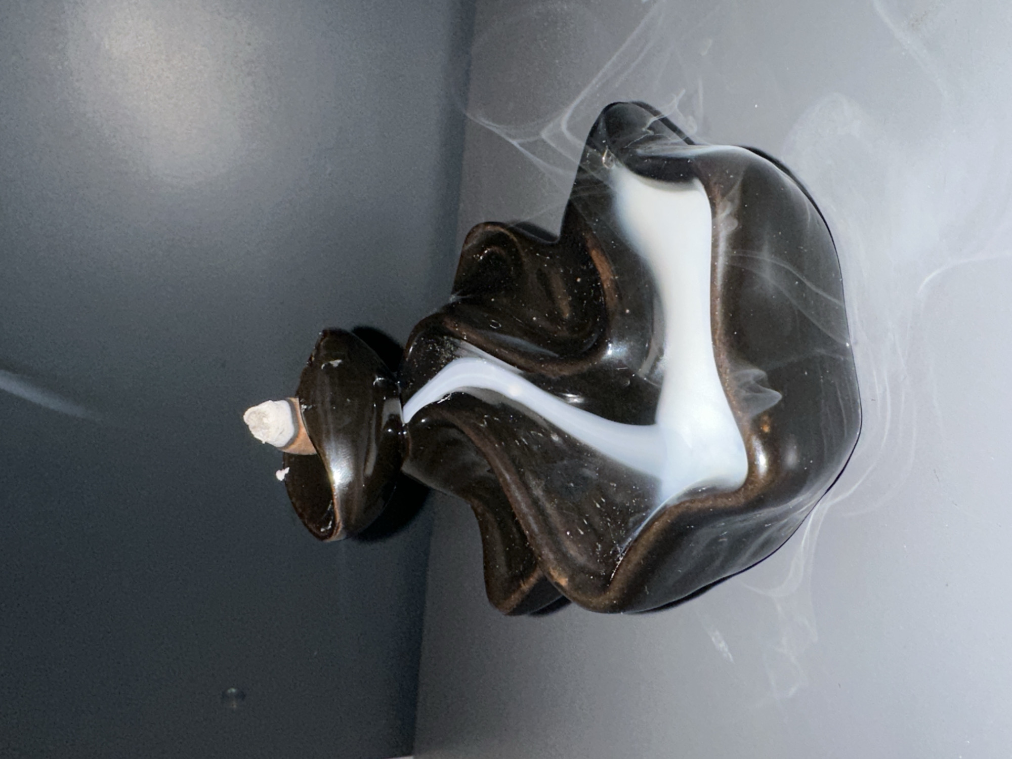 A brown and white incense burner is positioned in the corner of a room where two walls meet. Smoke emanates from the incense stick placed in the holder. The walls are painted in shades of gray. The location is not discernible from the image, and no information about Krasnoyarsk, Russia, can be inferred. There are no people visible in the image.