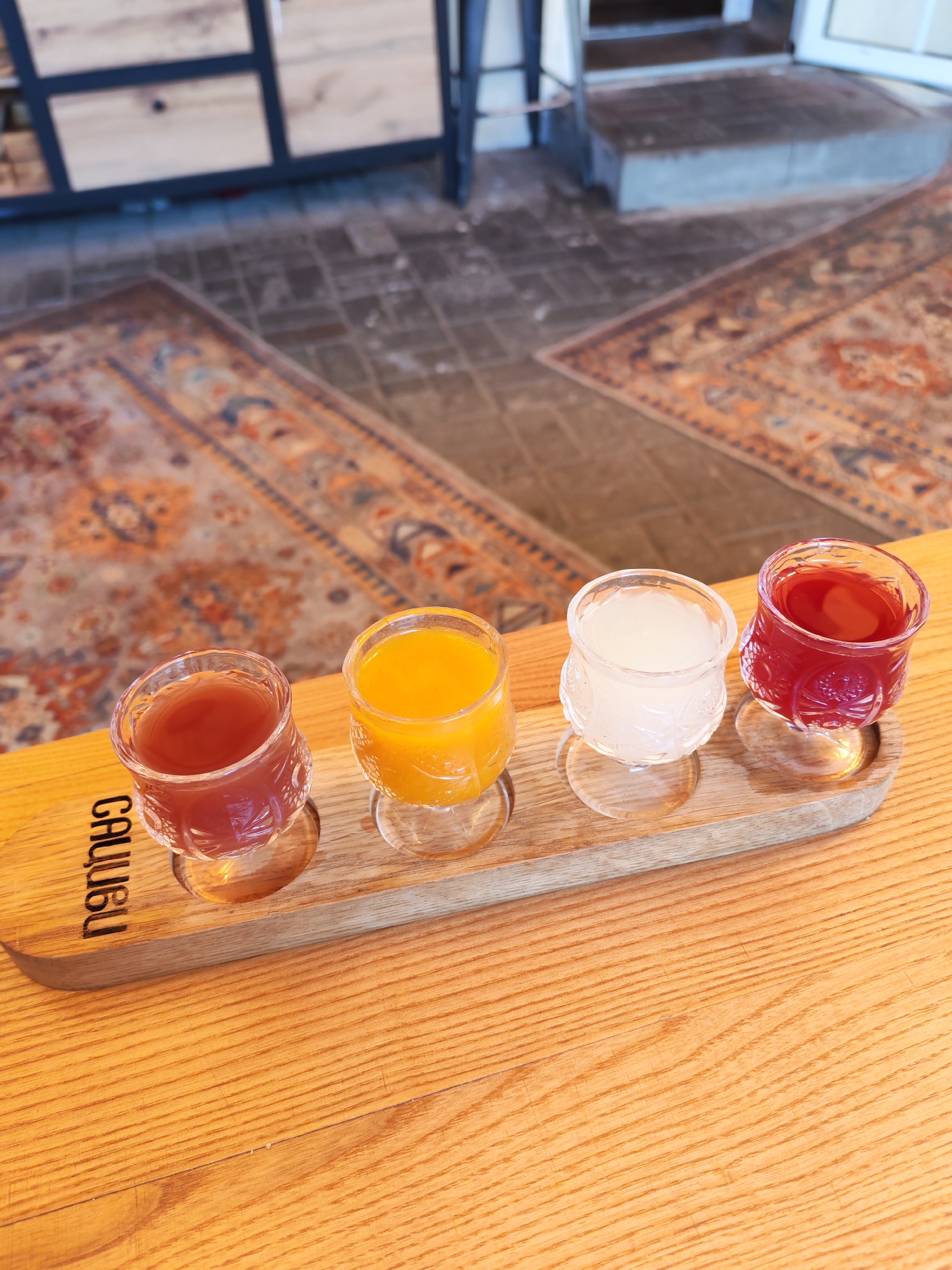 This is a close-up, eye-level, vertical, medium shot of a wooden tray with four small, clear glasses containing various colored beverages. The tray is resting on a wooden table. From left to right, the drinks appear to be a reddish-brown liquid, a bright orange liquid, a cloudy white liquid, and a deep red liquid. The glasses have a patterned texture on their lower halves and are sitting on small clear glass coasters. The tray has the word "CAULISU" engraved on its left side. In the background, out of focus, is a patterned rug on a brick-like floor and a piece of furniture with wooden panels and dark metal framing. The scene is indoors. There are no people visible. This image was taken in Bol'shie Mkhi, Russia.