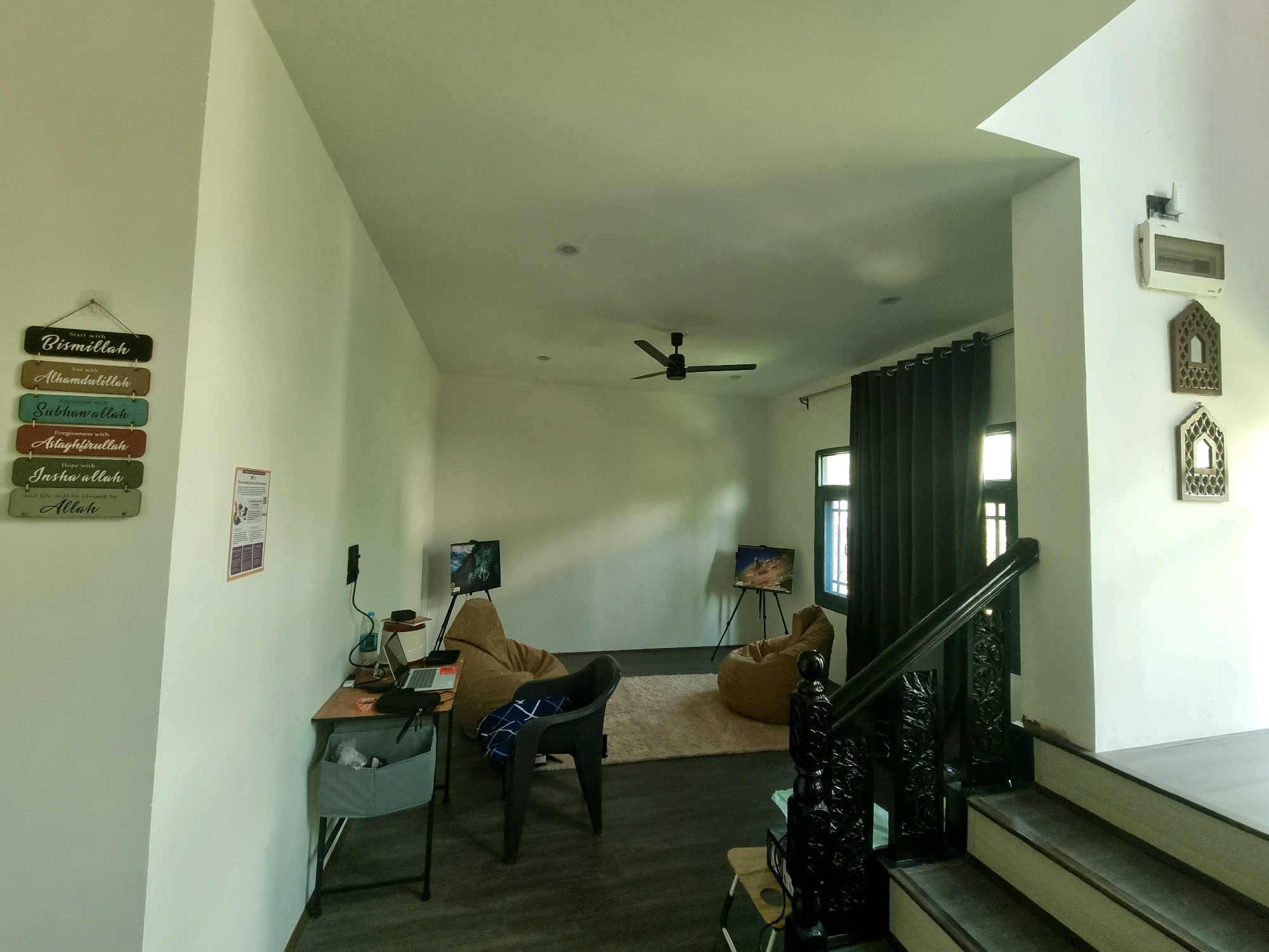 The image depicts an indoor scene, likely a living room or communal space. On the left wall, a decorative Islamic plaque with the text "Bismillah," "Alhamdulillah," "Subhanallah," "Astaghfirullah," and "Insha'allah" is hung. Adjacent to it, a small desk with a laptop, a bottle of water, and some papers is set up. Further into the room, two large beanbag chairs are placed on a rug, with small easels holding what appear to be digital art displays in front of them. A dark grey plastic chair is positioned between the beanbags. A ceiling fan is visible in the center of the room. On the right side of the image, a dark wooden staircase with intricate carvings leads upwards. A window with dark curtains is visible behind the stairs. On the wall near the stairs, a white electrical breaker box and two decorative wooden window frames are mounted. The walls are painted a light color, and the floor is dark. The environment appears to be a residential or common area.