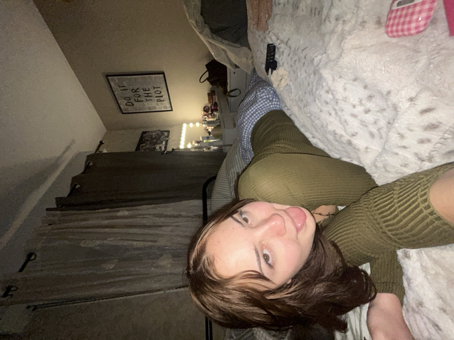 A young woman with brown hair and an olive green shirt lies on a plush, white blanket. She sticks out her tongue, facing the camera. A checkerboard sock sits near her. The room includes a bed with a grey-striped blanket, a shelf with fairy lights, and a framed print reading "Do it for the plot" hanging on the wall. A remote control is positioned on the white blanket. The image is taken from an awkward angle, possibly from a cell phone.