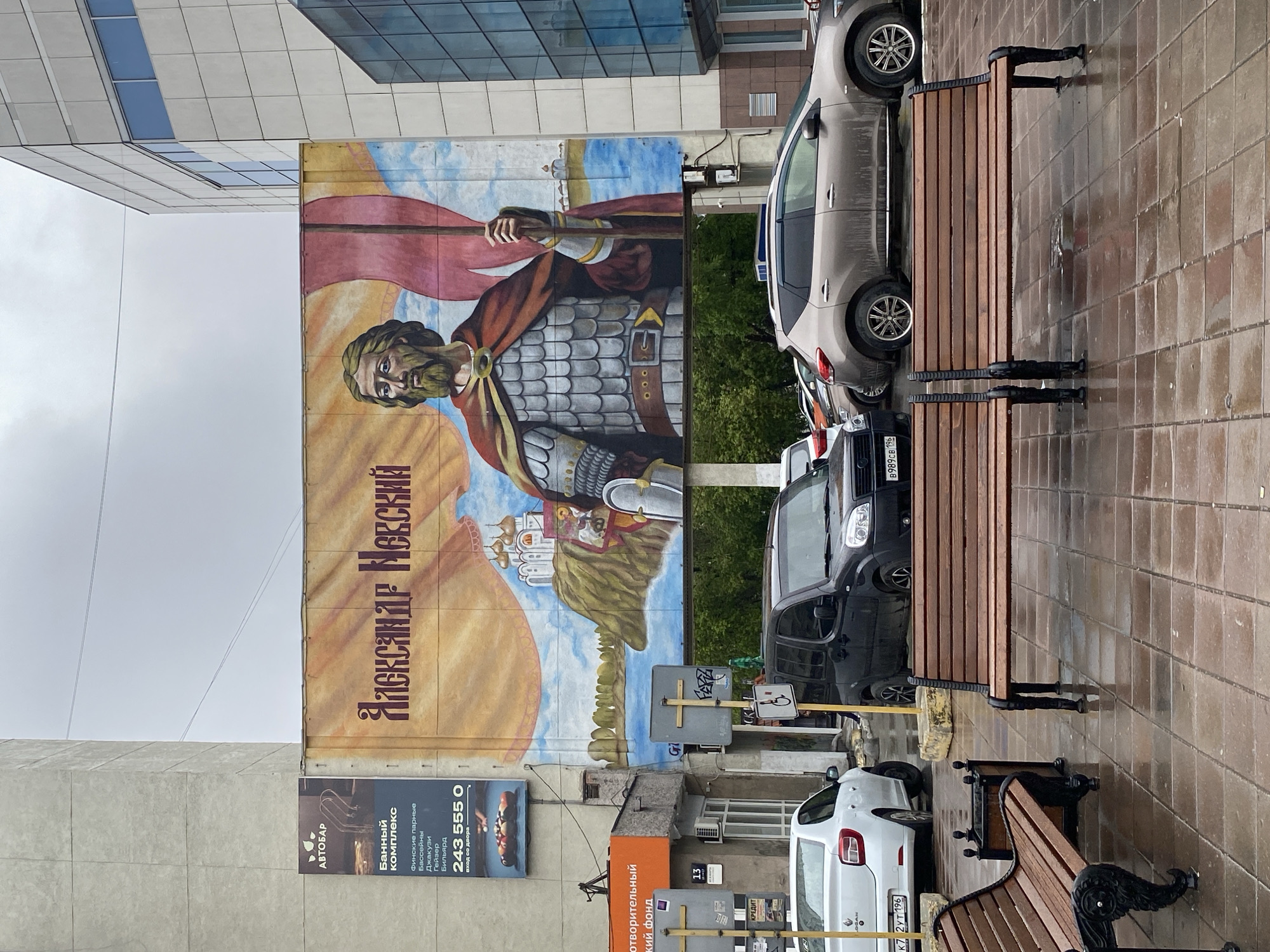 The image depicts a street scene in Yekaterinburg, Russia, featuring a large billboard mural of Alexander Nevsky. The billboard is positioned on the side of a building and is partially obscured by parked cars. There are several parked cars next to a bench that's on the sidewalk. Signage in Russian is visible near the billboard, including advertisements for a bathhouse complex and a credit organization. The weather appears to be overcast or rainy, as the sidewalk is wet.