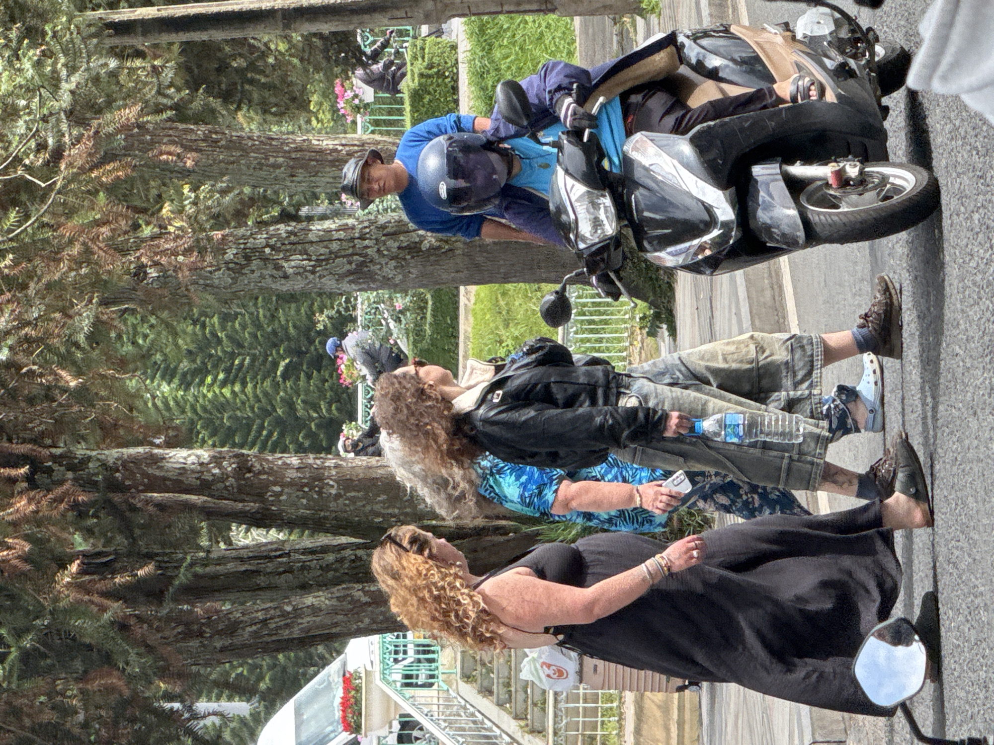 A medium shot shows a group of people on a street in Da Lat, Vietnam, with a woman in a long black dress walking to the left, followed by another woman in a blue patterned shirt and a third person in a black leather jacket. A person wearing a blue shirt and a helmet is riding a black and tan scooter in the foreground, with a man in a blue shirt and a hat standing behind them. The environment includes trees with sparse foliage, greenery, and a white vehicle in the background. A white plastic bag with a red logo is visible near the woman in the black dress. The ground is paved with asphalt.