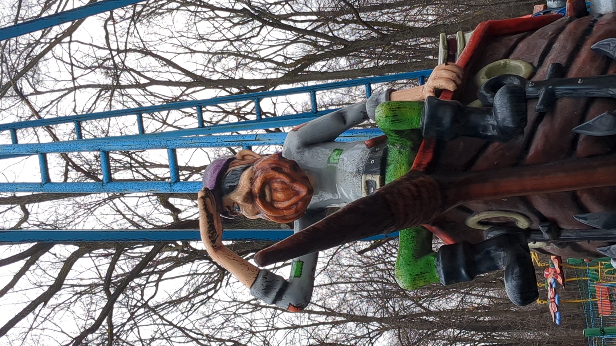 The image captures a distorted perspective of a pirate ship-themed amusement park ride in Ulyanovsk, Russia, likely taken from a low angle. The ride features a cartoonish pirate figure with an exaggerated nose, wearing a purple cap and a gray shirt with a belt. The pirate is holding onto the side of the ship. The ship itself is dark brown and red, adorned with black cannons. A blue metal frame supporting the ride is visible against a backdrop of bare tree branches and a gray sky. In the background, a brightly colored playground structure can be seen.