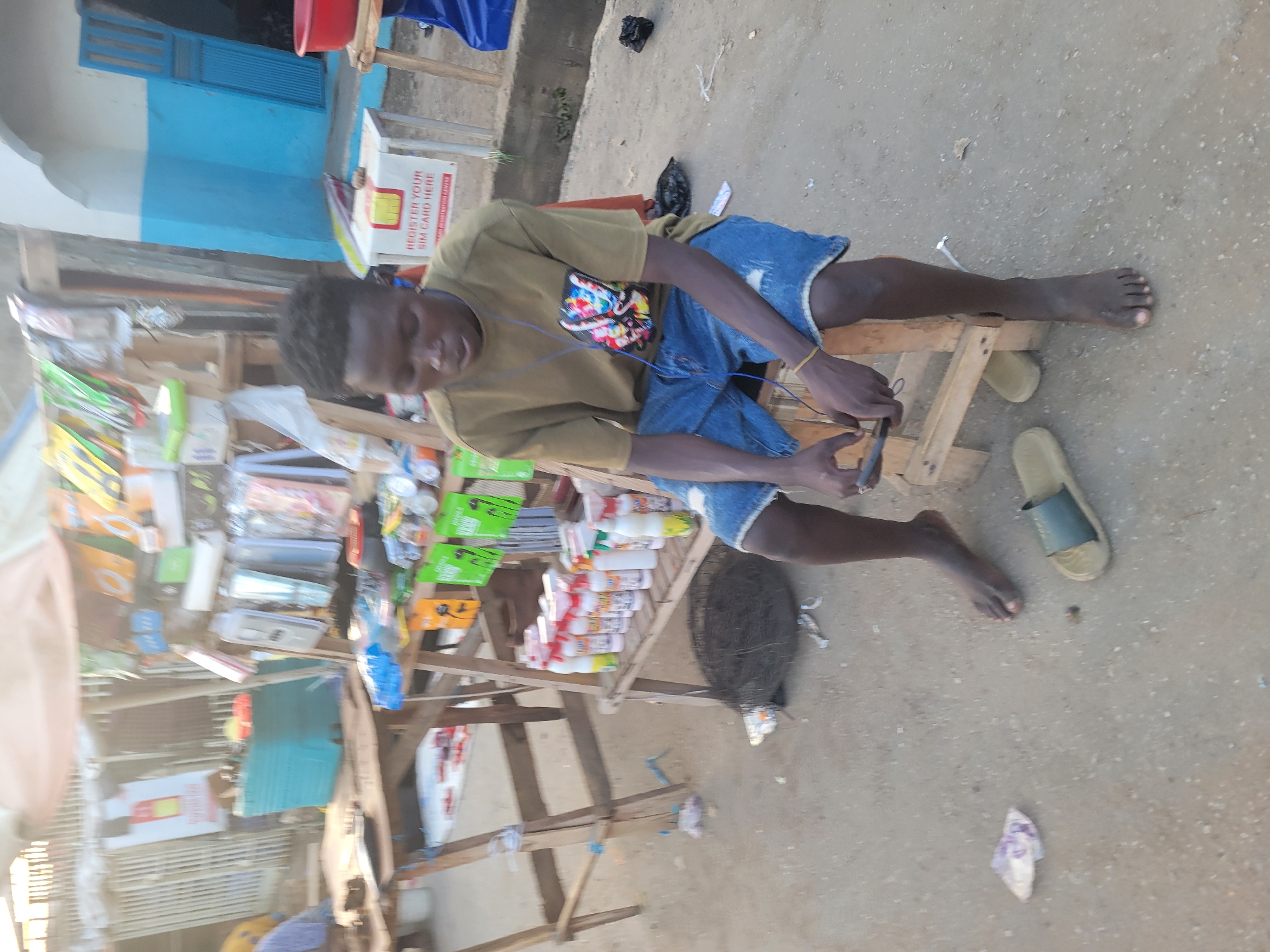 A young man is seated on a wooden structure in what appears to be an outdoor marketplace in Madalla, Nigeria. He is light-skinned, wearing a light-brown T-shirt, jean shorts, and has black hair. The man is barefoot and is looking down, seemingly focused on a mobile phone in his hands.
He is positioned next to a makeshift wooden stall stacked with various items including packages of goods, possibly for sale. A sign that reads "Register Your SIM Card Here" is visible on the stall. A woven basket sits next to the wooden structure the man is seated on.
The ground is a mix of dirt and concrete, with sandals and a piece of discarded trash scattered nearby. In the background, there is a building with a white and blue exterior.
