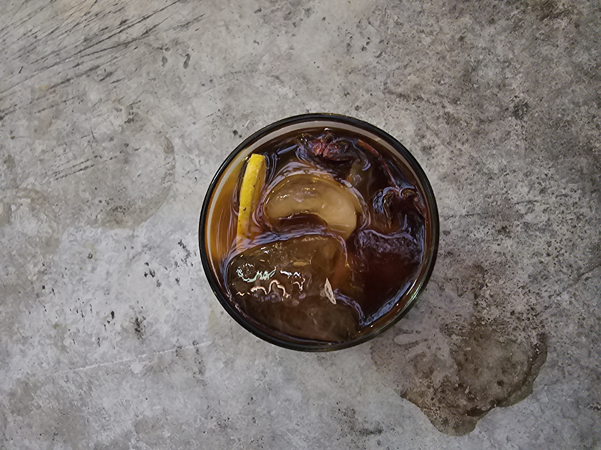A top-down, medium shot captures a glass of iced coffee with lemon on a gray, textured concrete surface in Yogyakarta, Indonesia. The glass, filled with dark brown liquid, ice cubes, and a slice of lemon, is positioned slightly off-center to the right. The concrete shows signs of wear and tear, with visible texture variations and a damp, darkened patch to the lower left of the glass, suggesting a spill. The lighting is even, highlighting the reflections on the ice and the liquid's rich color. No individuals are present in the frame.
