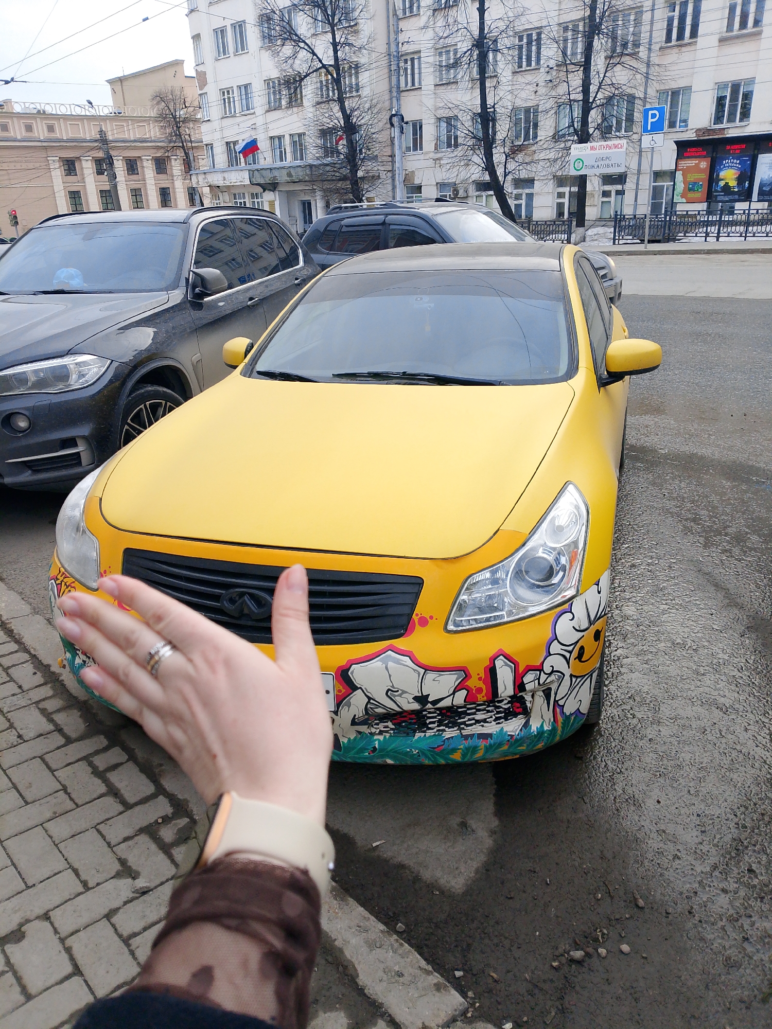 The image shows a yellow Infiniti car with graffiti on the front bumper parked on a city street. A person's hand is extended towards the car. Adjacent to the yellow car is a dark-colored SUV. In the background, there is a multi-story building with the Russian flag displayed. Further in the background on a pole is a sign with the letter "P" on it. The location is identified as Izhevsk, Russia, based on visual cues such as architectural style and signage. The weather appears overcast, and the street surface is wet.