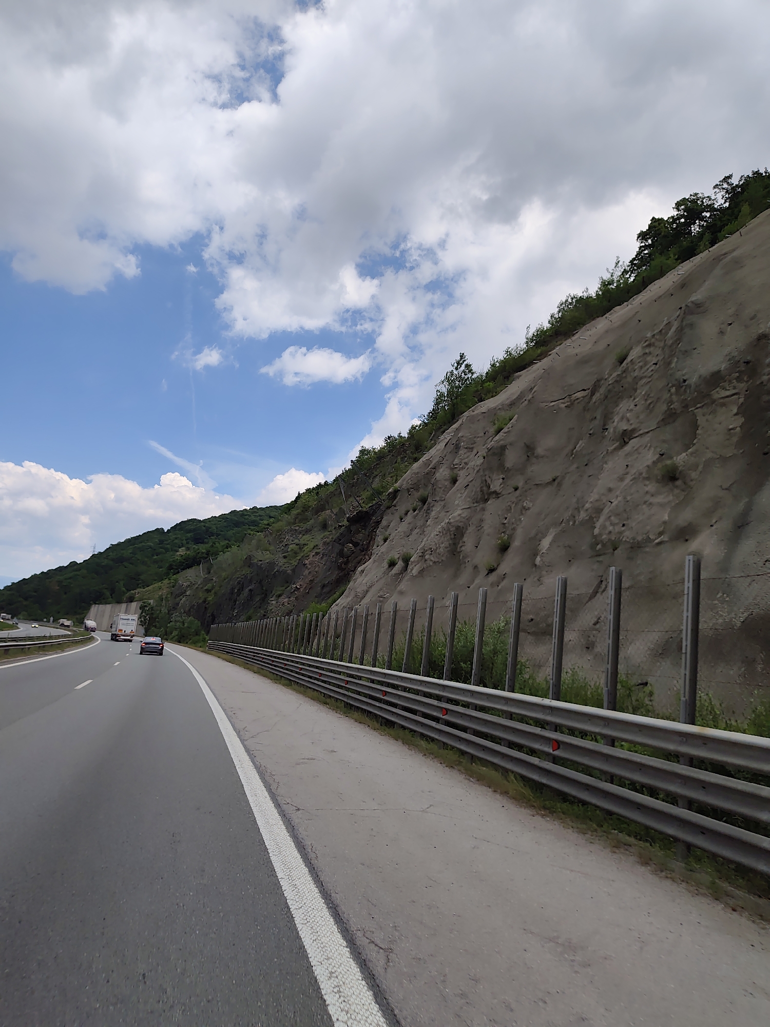 The image depicts a highway scene near Malo Buchino, Bulgaria. A multi-lane highway curves to the left, with cars visible in the distance. To the right of the highway, a steep, rocky hillside is reinforced with netting and vertical posts. A metal guardrail runs along the edge of the road. The sky is blue with scattered white clouds. The vegetation on the hillside is sparse, with some trees and bushes visible at the top.