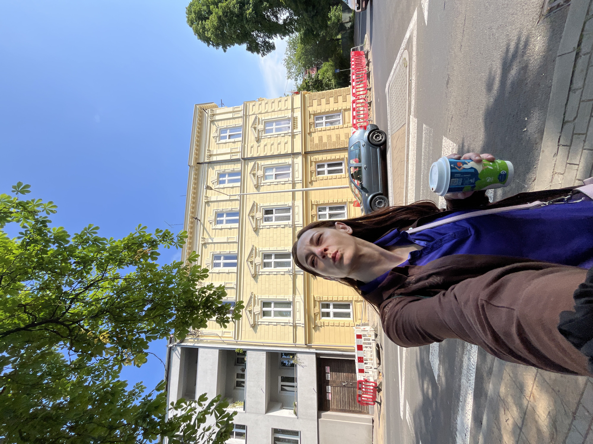 A woman is taking a selfie in an urban environment on a sunny day. She is holding a paper coffee cup in her right hand, and her left arm is extended. The woman is wearing a brown jacket over a purple shirt and has long brown hair. In the background, there are buildings, a street with a crosswalk, a car, and trees. The buildings appear to be of different architectural styles, with one being yellow with ornate trim and the other being gray and modern. There are also temporary red and white construction barriers visible on the side of the road. The sky is clear and blue.