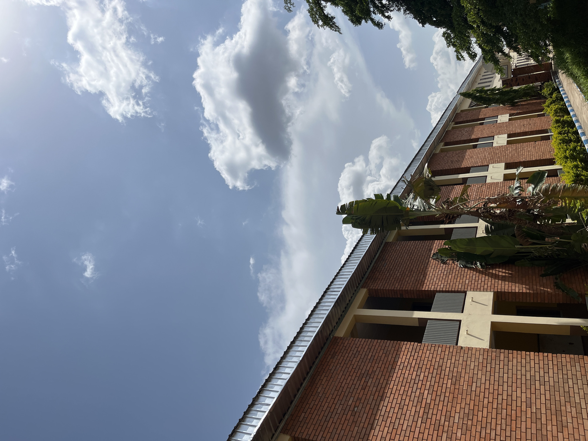 The media file shows a low-angle shot of a modern brick building under a partly cloudy sky. The building has multiple floors with a metallic roofline and several visible windows. Large, lush green leaves, likely from a tropical plant, hang over the top of the building. The sky is a clear blue with scattered white clouds, suggesting it is daytime. The image appears to be taken outdoors, possibly in a residential or commercial area. There is no visible text in the image. The lighting and cloud formations indicate it might be late morning or early afternoon. The setting appears to be a warm climate, consistent with Jos, Nigeria, as per the location context. The overall mood is serene and bright.