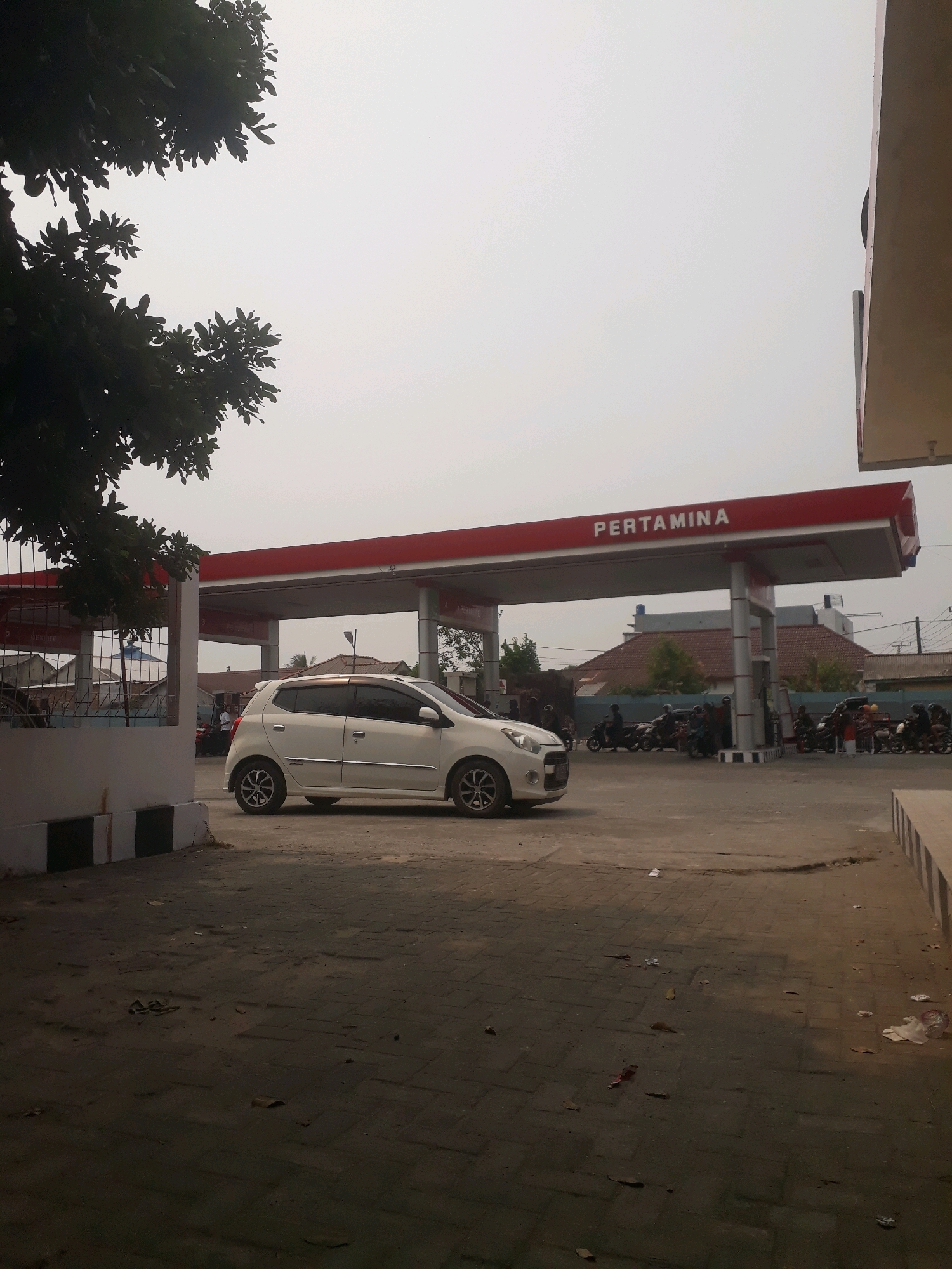The image depicts a scene at a Pertamina gas station in Pangkalpinang, Indonesia. A white car is parked on the asphalt in front of the gas station's canopy, which is marked with the "PERTAMINA" logo in red lettering. Several motorcycles are also visible, with people standing around them, suggesting they are either refueling or waiting. The environment appears to be a typical urban setting, with some buildings and trees in the background. The ground is paved with interlocking bricks, and there is some litter scattered on the ground, indicating a public space. The sky is overcast, suggesting a possibly hazy or cloudy day.