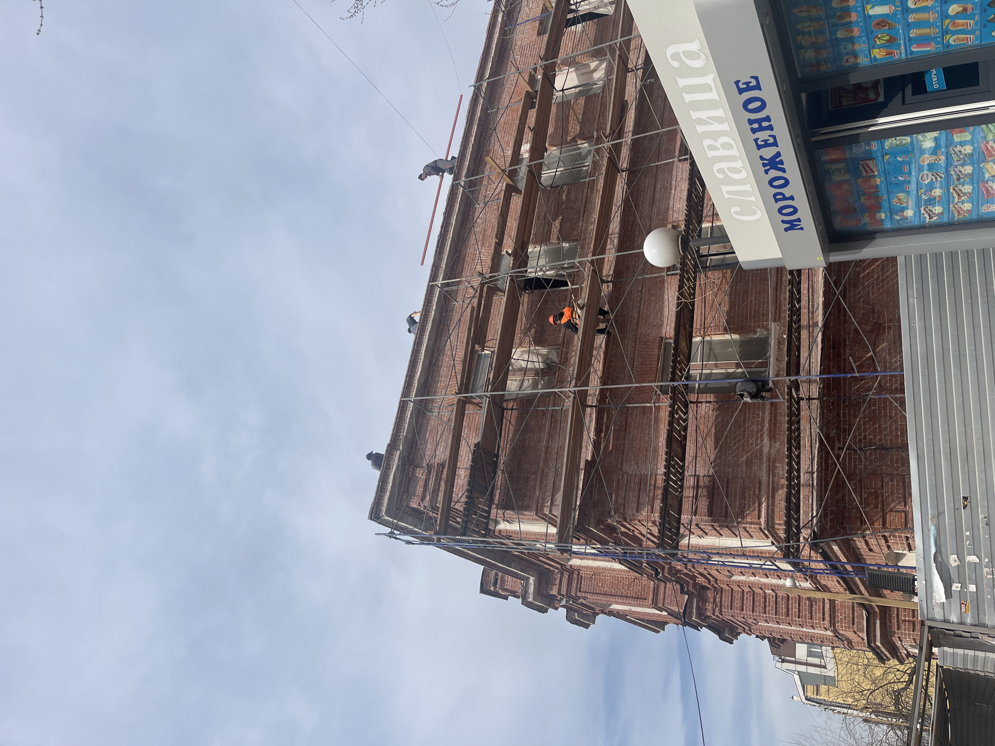 Here is a description of the image:
The image shows a red brick building in an urban environment, likely undergoing renovations. Scaffolding surrounds the building, and several workers are visible on the scaffolding at different levels. One worker is wearing an orange safety vest. There is a blue and white sign on the side of the building, which is written in a Cyrillic script that says "СЛАВИЦА МОРОЖЕНОЕ", translating to "Slavitsa Ice Cream." Next to the building is a display with numerous ice cream products. The sky is overcast with a bluish-gray color, indicating daytime. There are a few visible buildings in the distance, but the focus is on the brick building and the ongoing construction or restoration work.