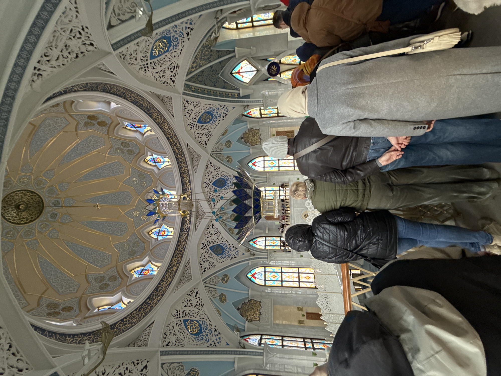 The image captures the interior of the Qolsharif Mosque, located in Kazan, Russia. Several people are present, appearing to be engaged in prayer. The focal point is the mosque's dome, adorned with intricate Islamic geometric patterns, stained glass windows, and decorative calligraphy. A chandelier hangs from the center of the dome. The architecture features detailed white arches and columns, contributing to the mosque's grandeur.