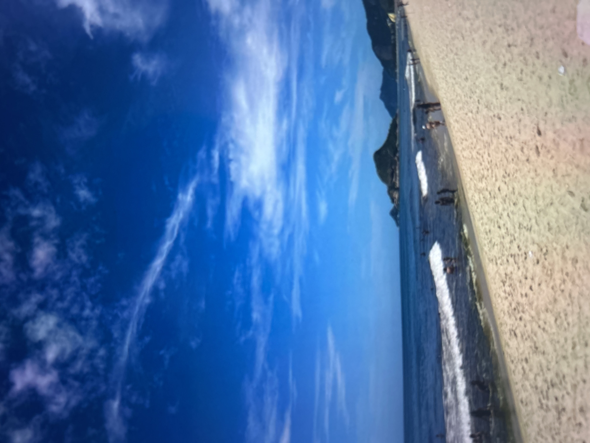 This media file shows a beach scene with a clear blue sky dotted with wispy white clouds. The ocean is visible with gentle waves approaching the shore. The beach itself is sandy and appears to be quite wide. There are several people on the beach and in the water. Some people are walking along the shoreline, while others seem to be wading or swimming in the ocean. In the background, a mountainous landscape is visible, suggesting a coastal location with rugged terrain. The lighting indicates it is daytime, likely sunny given the clear sky. The overall impression is one of a relaxed and pleasant day at the beach. No visible text is present in the image.