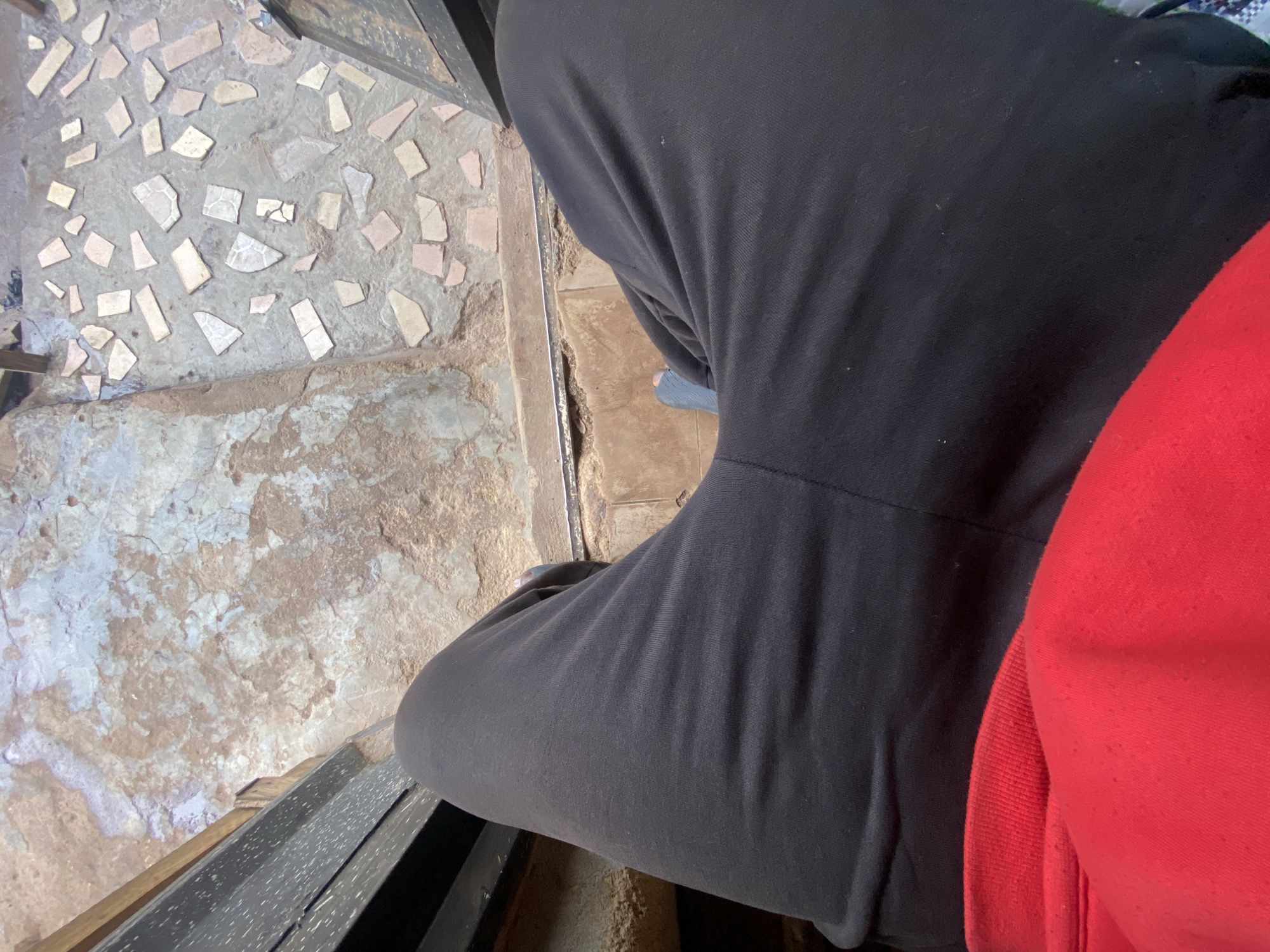 A person is sitting on a bench outdoors. They are wearing black sweatpants and a red hoodie. The ground is made of concrete and tile, with some dirt. The scene appears to be in a courtyard or patio area. The camera angle is from above, looking down at the person's legs and the ground.