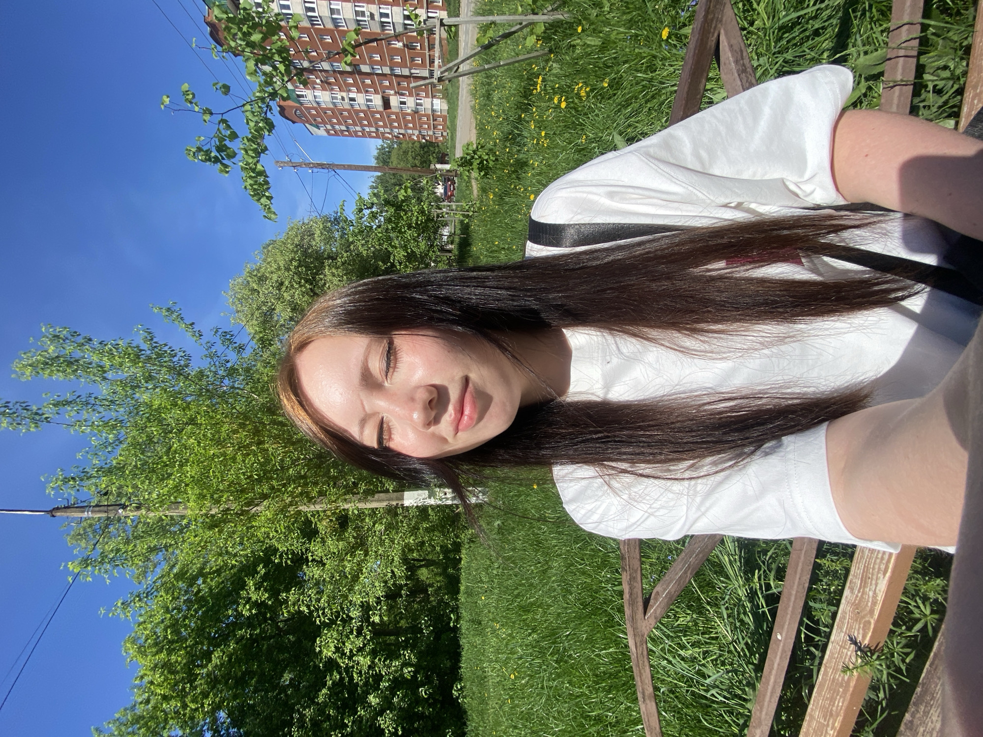 A young woman with long, dark brown hair is sitting on a wooden bench outdoors on a sunny day. She is wearing a white t-shirt and a black bag strap is visible over her shoulder. Her eyes are closed and she appears relaxed. Behind her is a grassy area with yellow flowers and trees, with a multi-story brick building in the background. The location appears to be in an urban park.