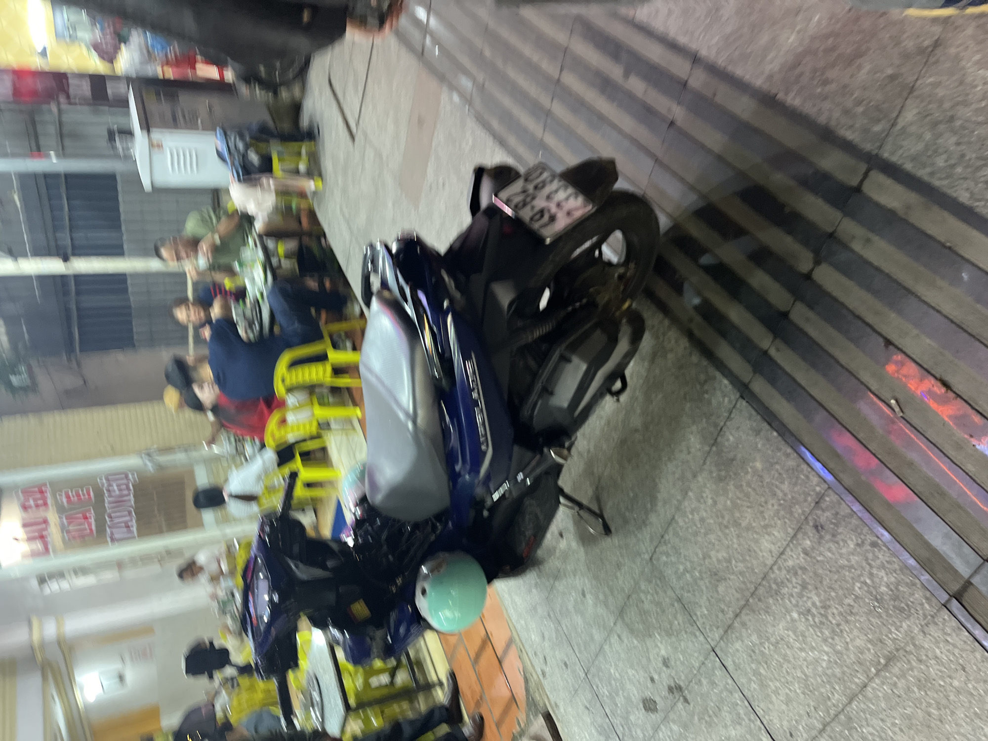 The image depicts a street scene in Da Lat, Vietnam, likely in the evening, with a focus on a parked dark blue motorcycle. The motorcycle has a light blue helmet attached to its left side. In the background, several people are seated at tables, some appearing to be dining. Yellow plastic chairs are visible around the tables. A sign with Vietnamese text in red is mounted on a building in the background. The ground is paved with square tiles and has a section with raised tactile paving for the visually impaired. The overall impression is a bustling street-side eatery.