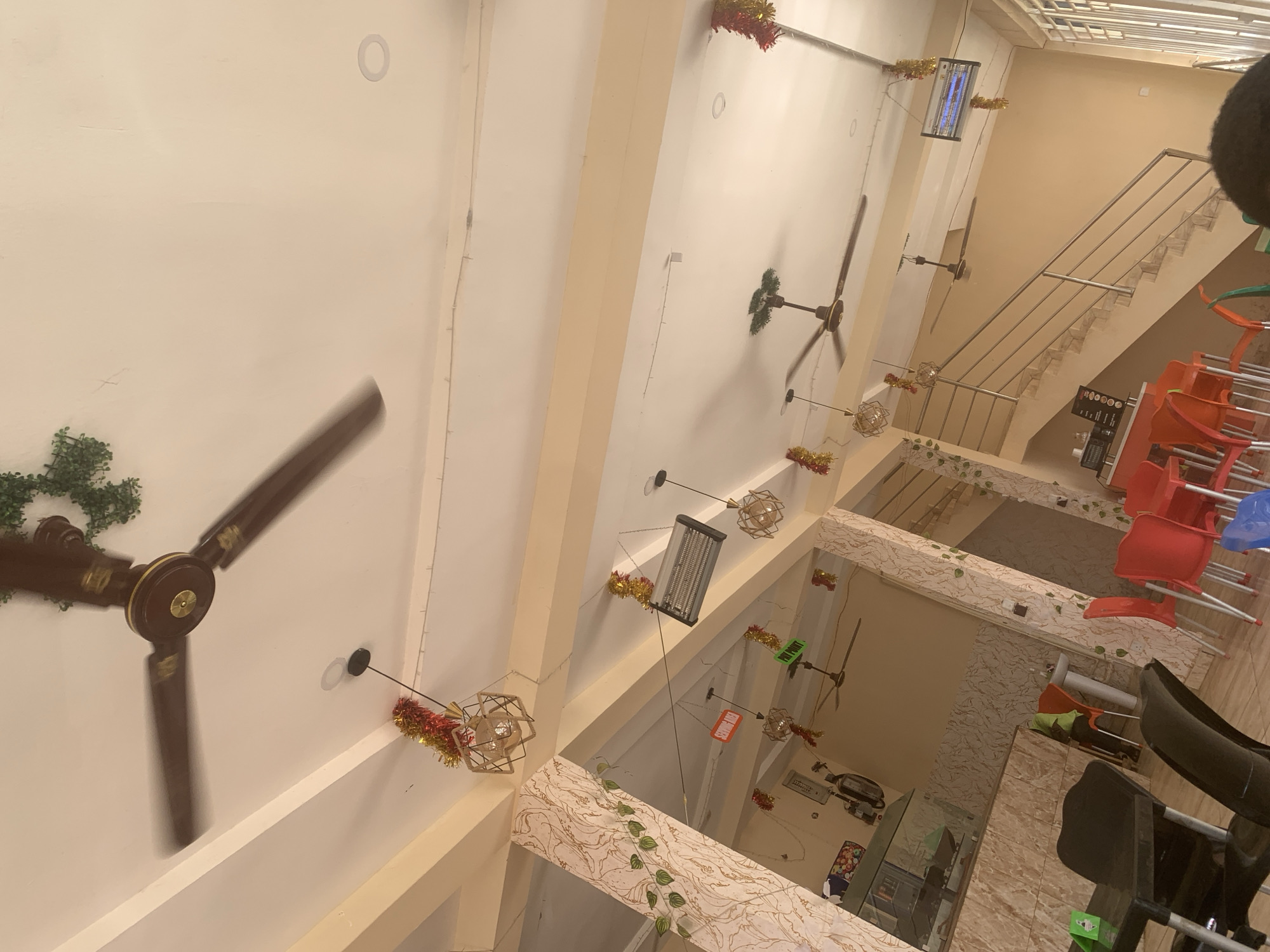 The image shows the interior of a building in Ungwan Liman, Nigeria. The space appears to be a commercial establishment, possibly a restaurant or cafe. The scene is captured from a high angle, looking down. There are multiple ceiling fans with brown blades, and some are decorated with green foliage or festive tinsel. The walls are light-colored. There are multiple stairs in the building. Tables and chairs are visible, with chairs stacked on some tables. There is a mix of seating colors, including red, blue, and green. There is a mirror with some decor on it.