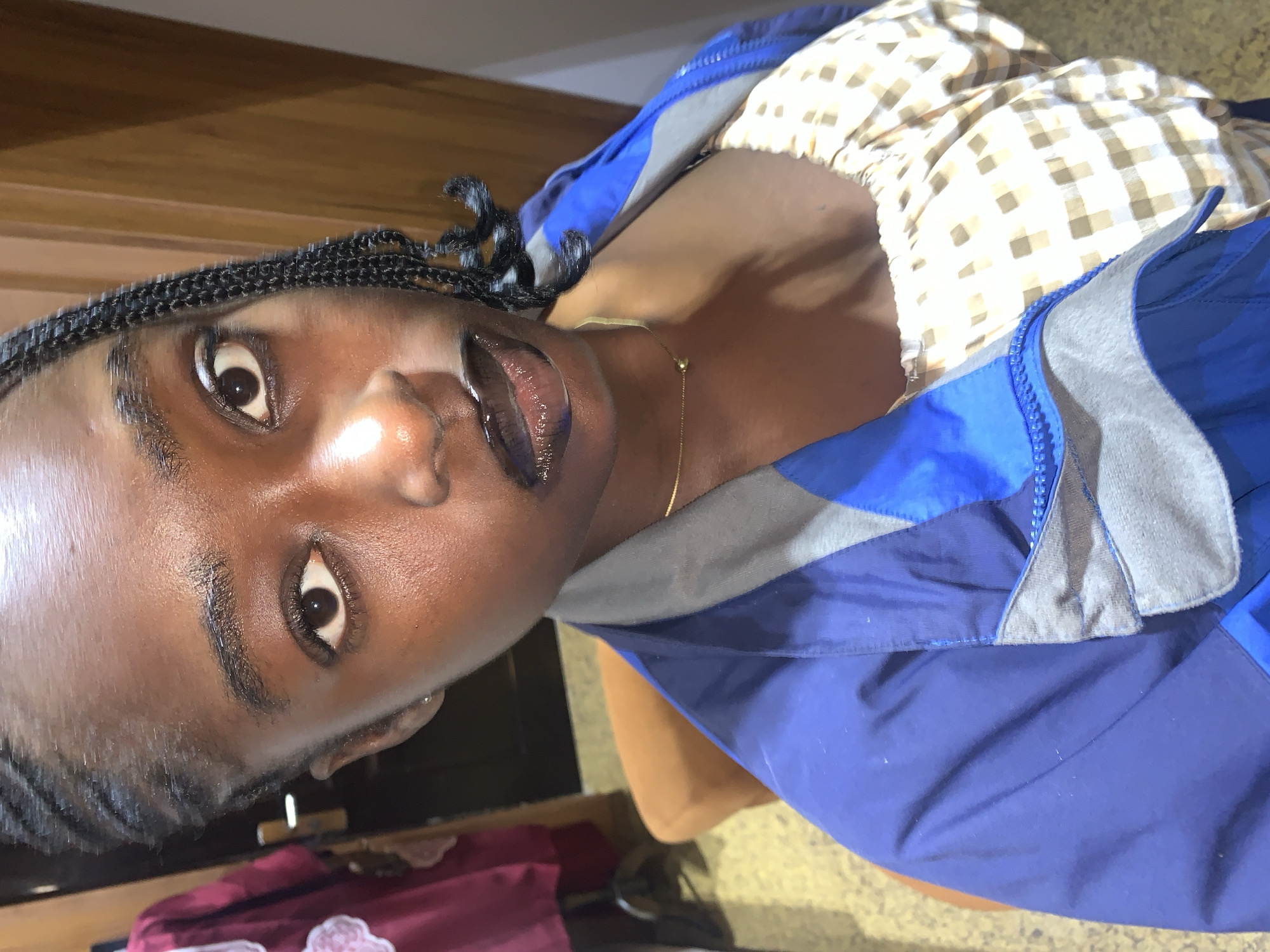 A close-up, waist-up, indoor selfie of a young Black woman with dark braided hair. She is wearing a blue and gray zip-up jacket over a cream-colored, gingham-print top. Her lips are painted with dark lipstick. She has a gold chain necklace with a small round pendant. The background appears to be a room with a wooden door and a piece of clothing on a rack to the left. The lighting is warm and highlights the sheen on her skin. The scene does not depict any recognizable landmarks or indicators of the specific location within Gura Top, Nigeria.