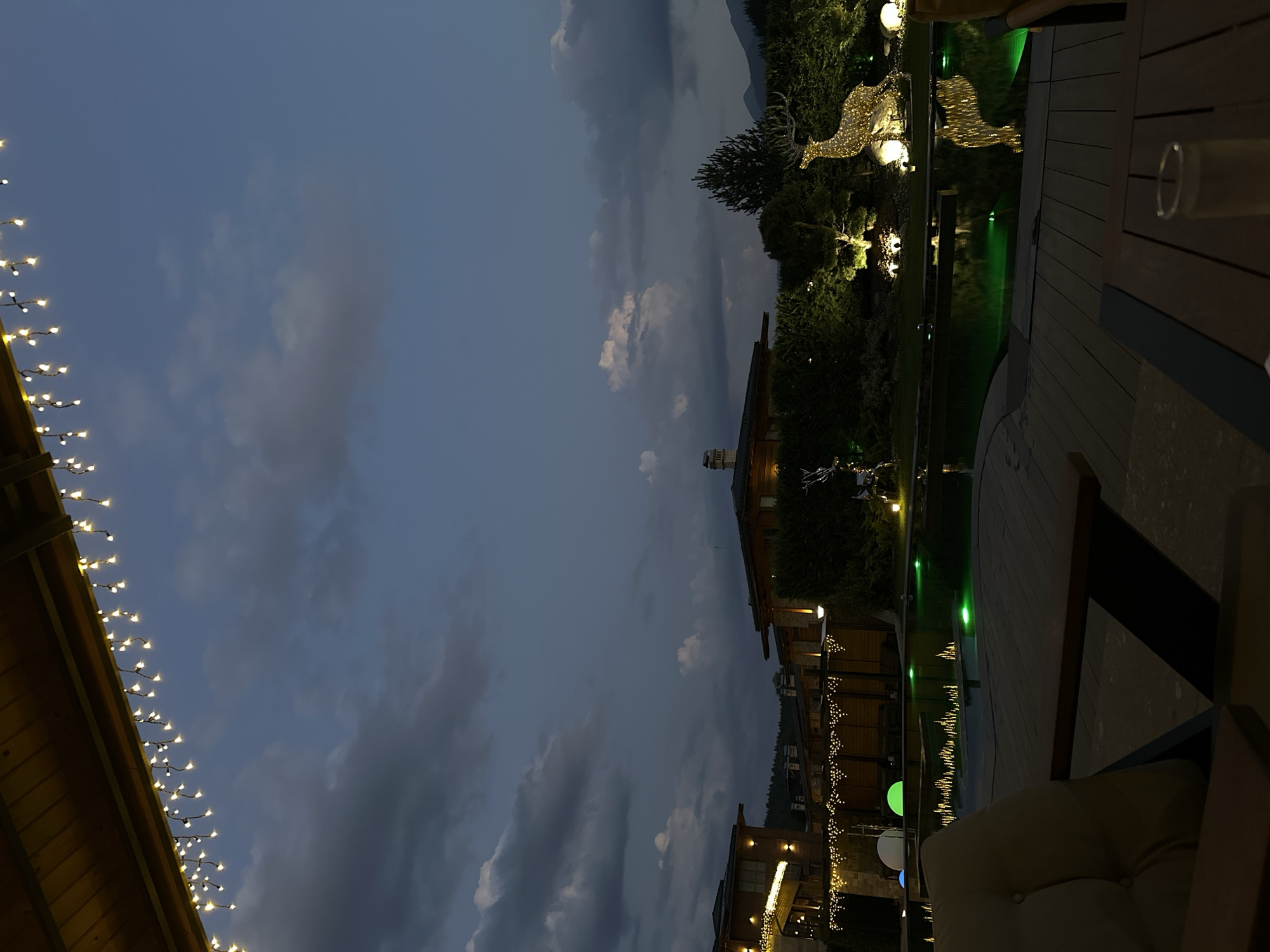 The image depicts an outdoor scene at dusk, likely at a resort or private residence in the city of Bania, Bulgaria. In the foreground, a wooden deck with a table and chair is visible, with a glass of liquid on the table. The deck overlooks a dark, rectangular swimming pool illuminated with green lights.
Beyond the pool, a landscaped garden features illuminated decorative elements, including a large, light-covered reindeer sculpture. Several buildings with wooden exteriors and lit windows are situated behind the garden, suggesting accommodations or a lodge. A string of fairy lights hangs from the eaves of a structure in the upper left corner. The sky is a deep blue with scattered clouds, indicating the transition from day to night. The overall atmosphere is serene and festive, with the lighting contributing to a welcoming ambiance.