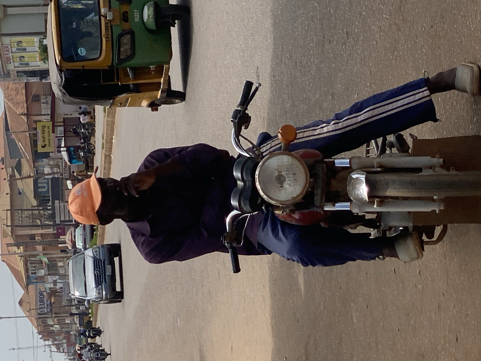 In Sabon Tasha, Nigeria, a dark-skinned man wearing an orange baseball cap and a purple shirt is seated on a red and chrome motorcycle. He is wearing dark blue pants with white stripes. The motorcycle has a prominent headlight and handlebars. The man is looking to his left and has his right hand on his chin. The background shows a street with other vehicles, including a silver SUV, a yellow and green auto-rickshaw, and several other motorcycles. Buildings line the street, some with signage. The ground is paved.