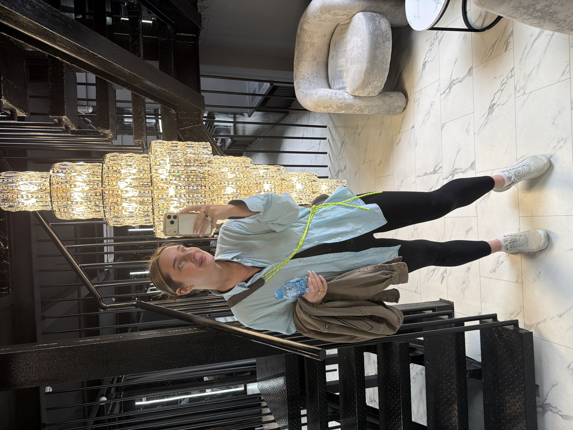 A woman stands on black metal stairs beneath a multi-tiered crystal chandelier, taking a selfie with her phone. She wears black leggings, white sneakers, and a light blue button-down shirt with a neon green strap across her chest. In her hand, she holds a bottle of water and a brown jacket. The stairs are next to a marble-tiled floor with a modern, rounded armchair nearby. The location is Moscow, Russia.