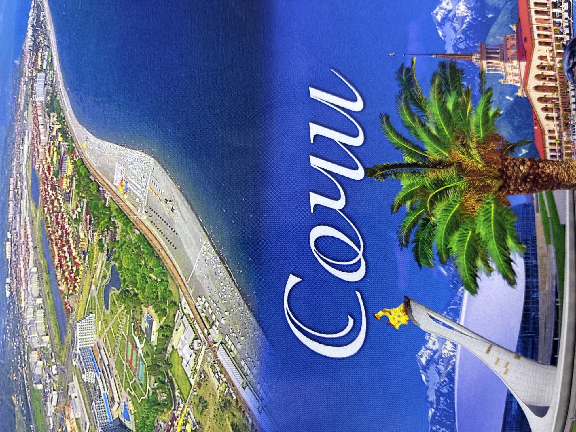 This image is a collage depicting different scenes of Sochi, Russia, not Abaza. The top portion shows an aerial view of a coastal city with buildings, green spaces, a beach, and the sea. The bottom portion features a close-up of a palm tree in front of snow-capped mountains and a white Olympic torch sculpture with a flame. To the right of the palm tree is a building with a spire, resembling a train station, and a dolphin sculpture. The word "Sochi" is written in large white cursive letters across the blue sky in the lower half of the image. There are no people visible in the frame.