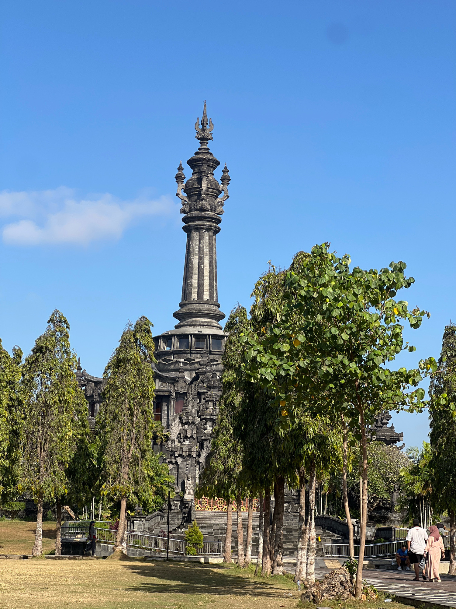 The image shows the Bajra Sandhi Monument in Denpasar, Bali, Indonesia. It is a tall, gray stone monument with intricate carvings and a spire at the top. The monument is surrounded by lush green trees and a clear blue sky. In the foreground, there is a grassy lawn and a paved walkway. Several people are visible in the scene, including a couple walking on the path and another person sitting on a bench. The overall atmosphere is peaceful and serene, indicative of a public park or historical site.